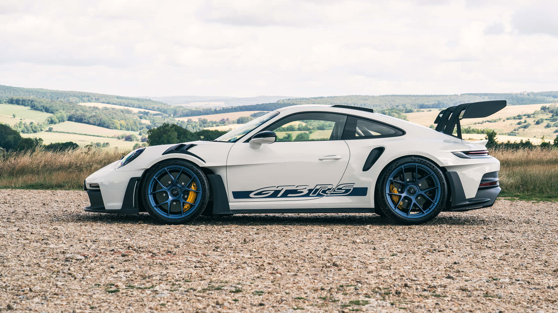  壁紙 — 一輛白色保時捷911 GT3 RS，配備藍色輪圈和打開的後尾翼，停泊在碎石路上，俯瞰著多雲天空下的連綿綠色山丘。