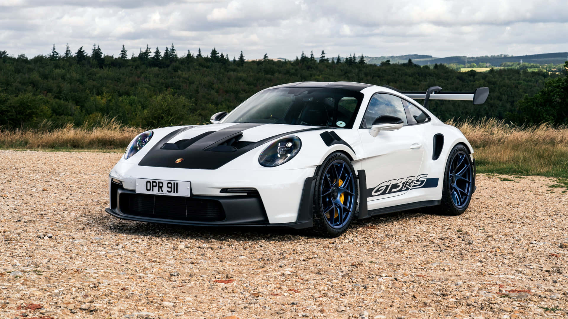  壁紙 — 一輛白色保時捷911 GT3 RS，帶有黑色賽車條紋和藍色輪圈，停放在碎石地上，背景是森林覆蓋的山坡，天空多雲。