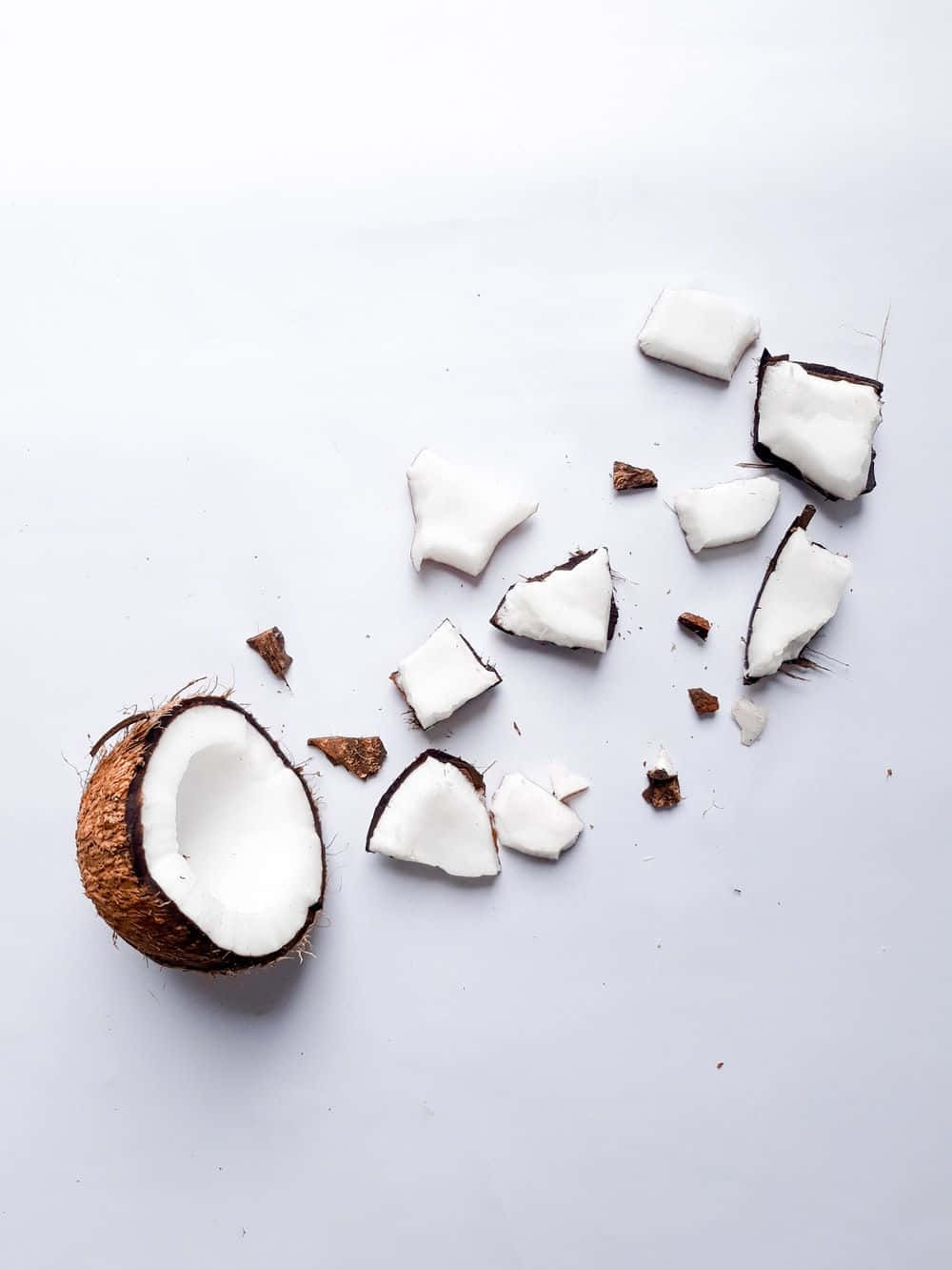 Coconut 桌布 壁紙 — 半個椰子，白色果肉碎片和棕色外殼碎片散落在乾淨的白色表面上。
