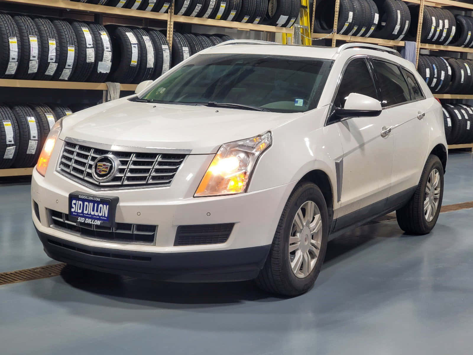 Cadillac Srx 桌布 壁紙 — 一輛白色Cadillac SRX SUV亮著車頭燈停放在輪胎倉庫，後面有輪胎架。