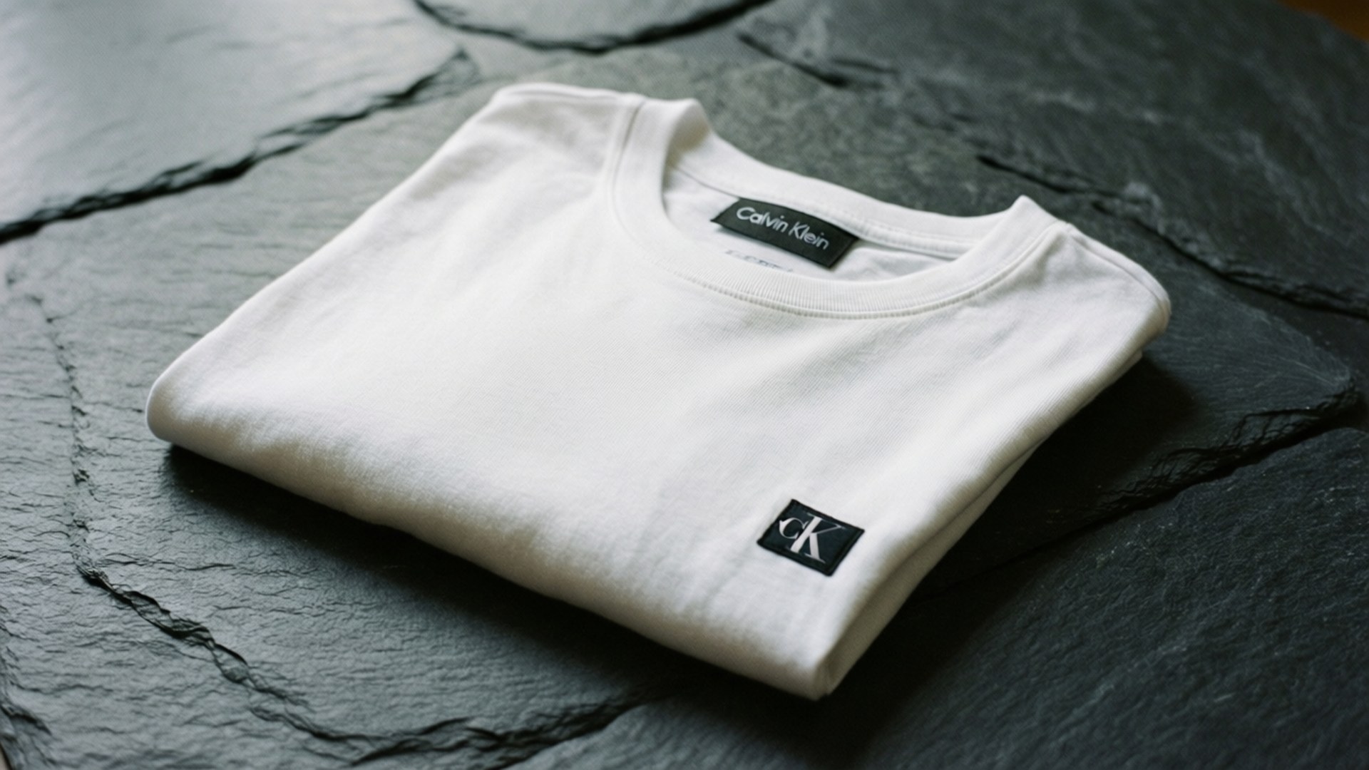 Folded Tee 壁紙 — 一件純白 Calvin Klein T 恤整齊折疊在深色紋理石板表面上。