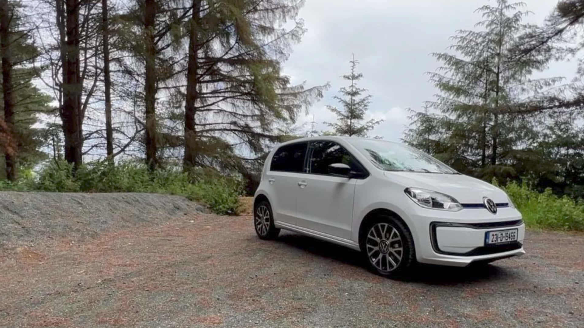  壁紙 — 一輛白色福斯 e-Golf 停泊在碎石路上，周圍是高聳的常綠樹，天空多雲。