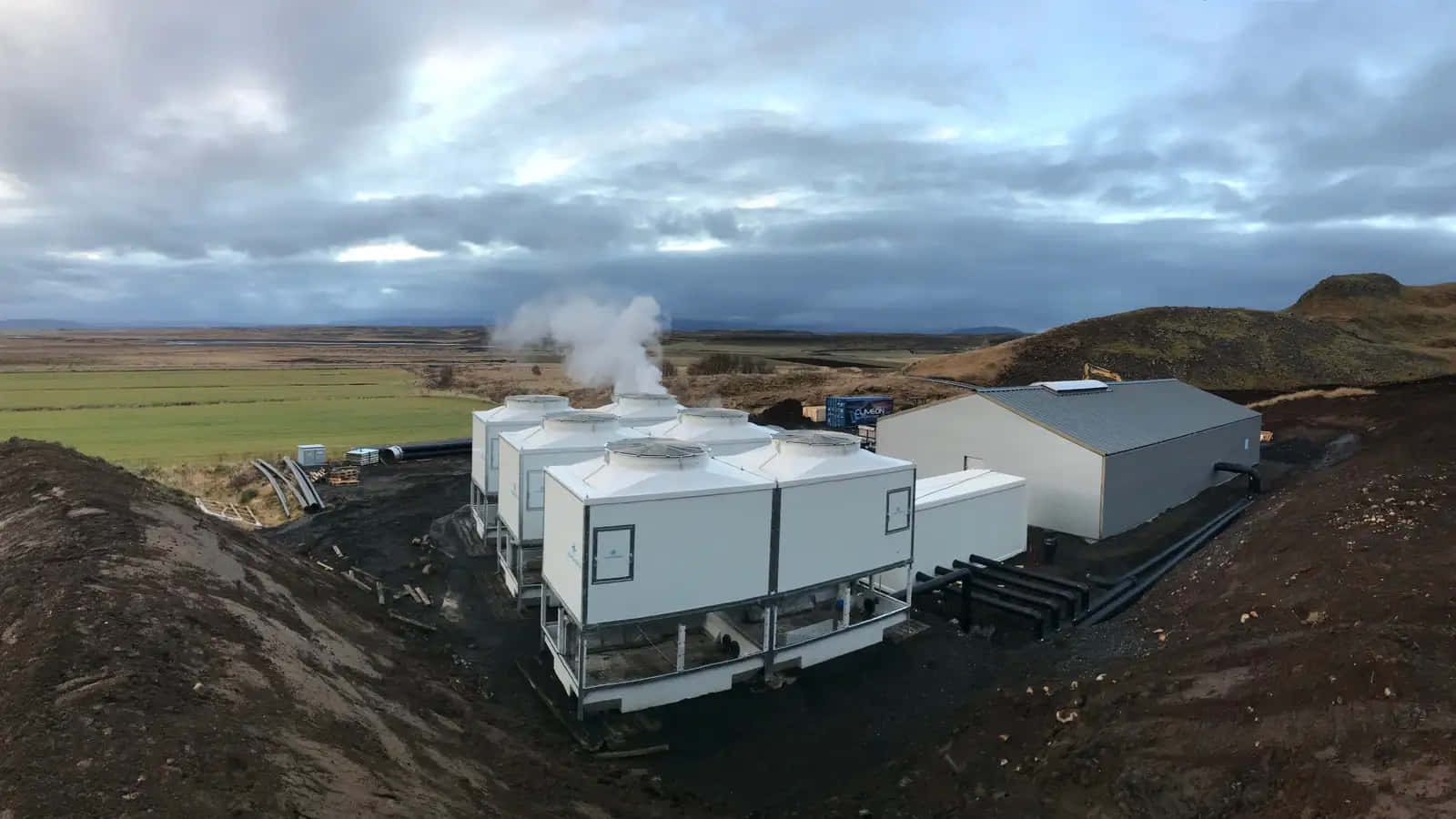  壁紙 — 白色工業地熱發電廠在陰天背景下，於崎嶇地形和綠色田野上方排放蒸汽。