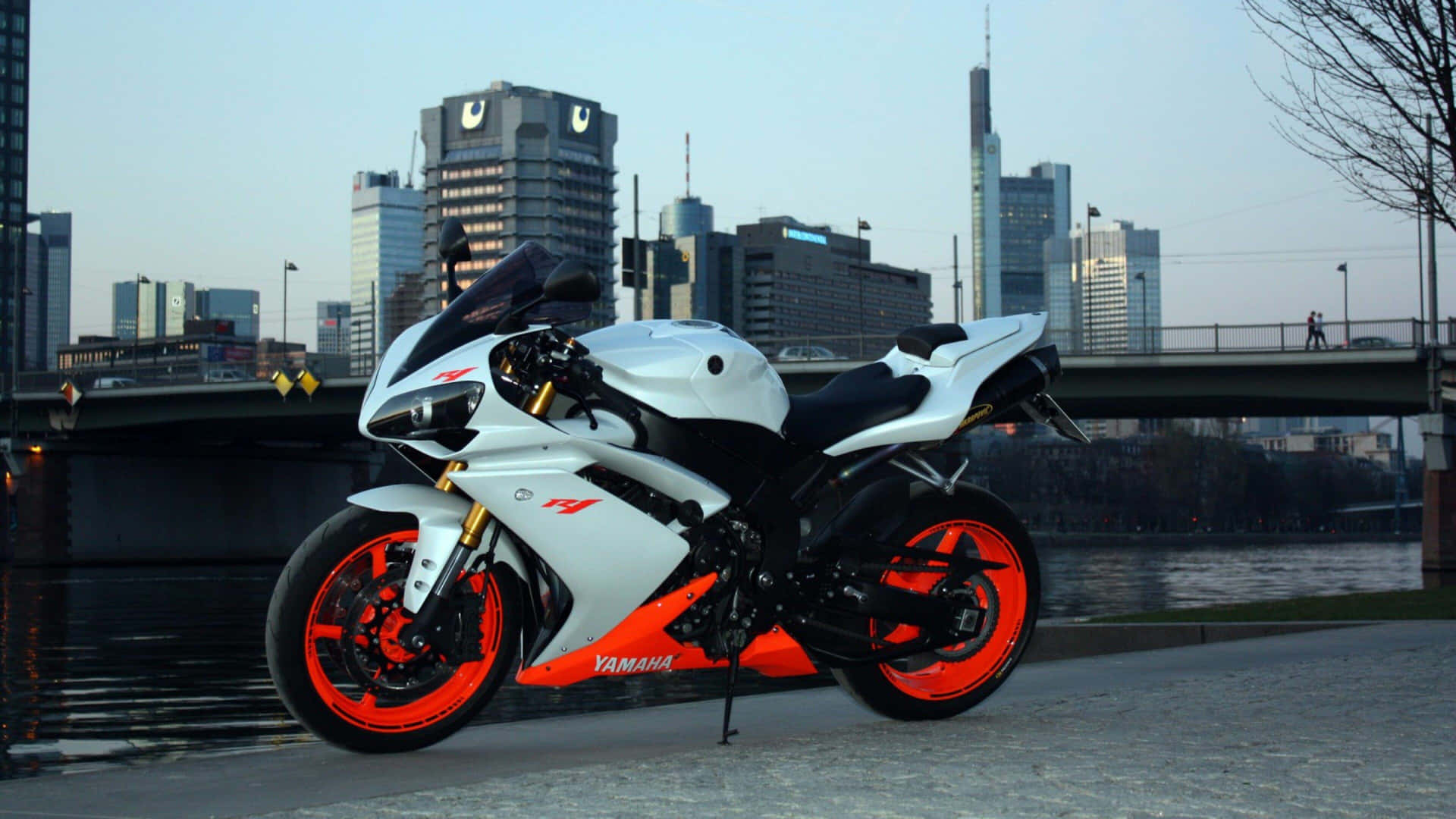  壁紙 — 一輛白色和橙色的Yamaha運動機車停泊在河邊小徑上，背景是黃昏時分的現代城市天際線。