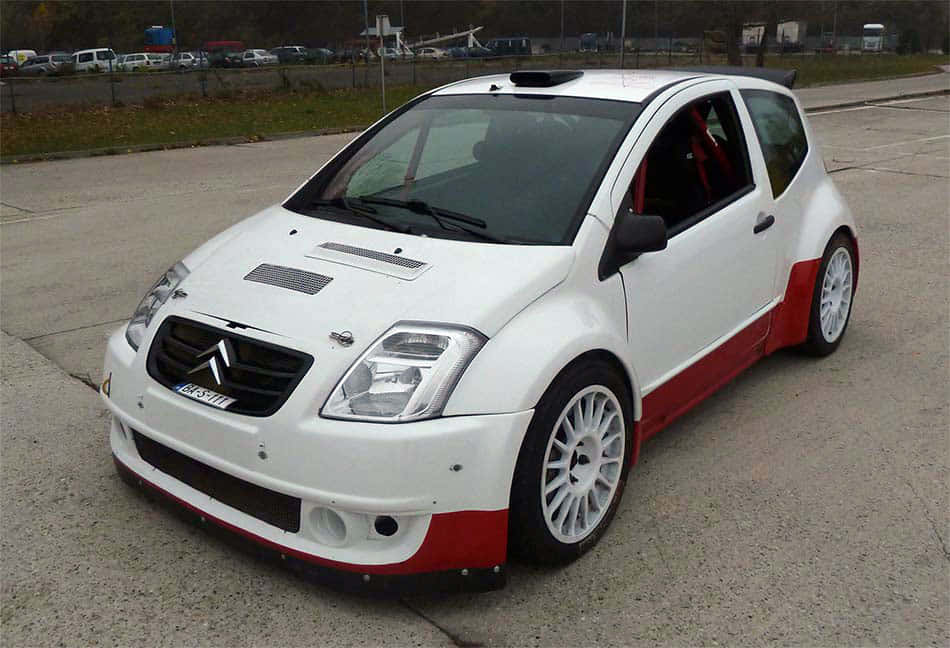Citroen C2 Super 1600 桌布 壁紙 — 一輛白色和紅色改裝的雪鐵龍C3拉力賽車停在有草地背景的混凝土場地上。