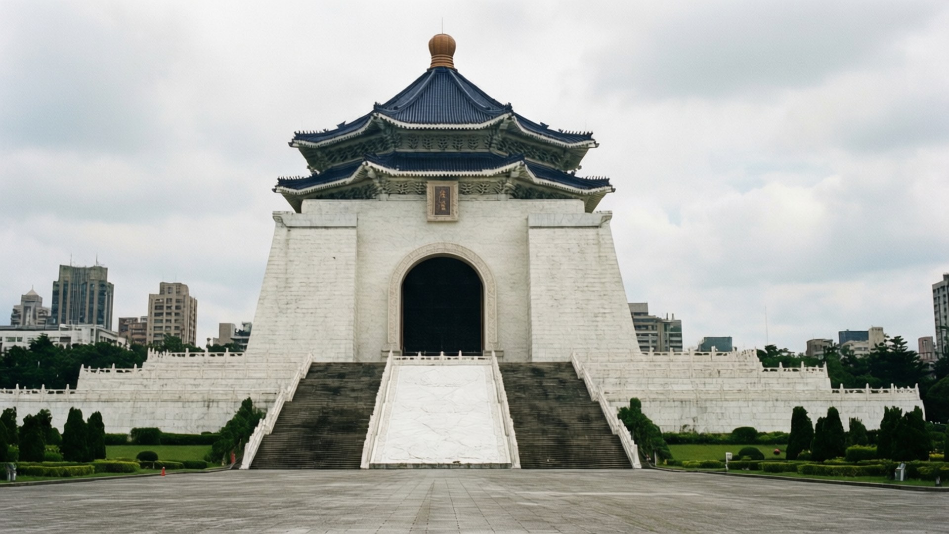 台灣 桌布 壁紙 — 白色宏偉的中正紀念堂，藍色琉璃瓦屋頂，在陰天之下，聳立於修剪整齊的花園和城市天際線之間。