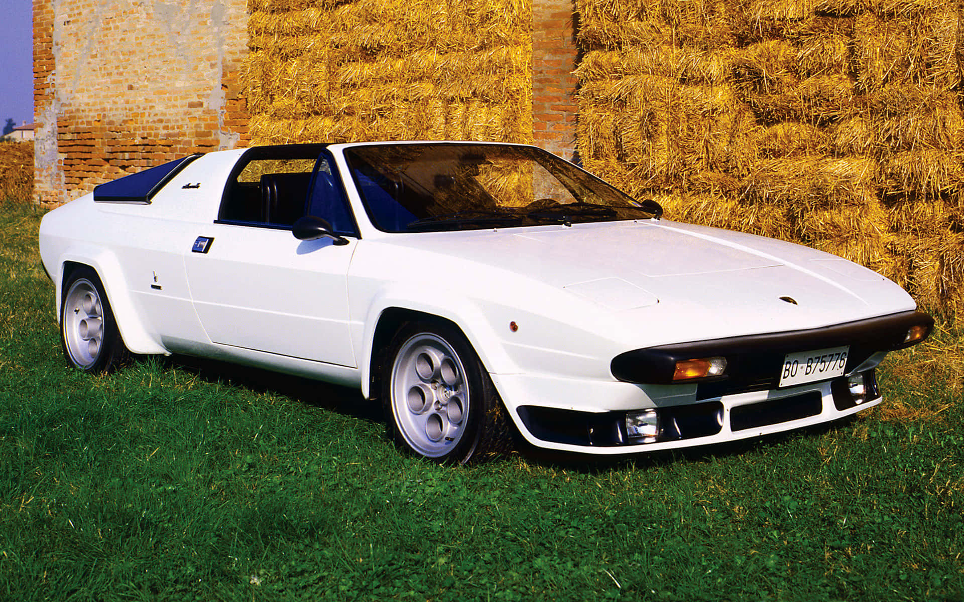 Lamborghini Silhouette 桌布 壁紙 — 一輛純白色的藍寶堅尼 Jalpa 跑車停放在翠綠的草地上，旁邊是質樸的磚牆和金黃色的乾草捆。