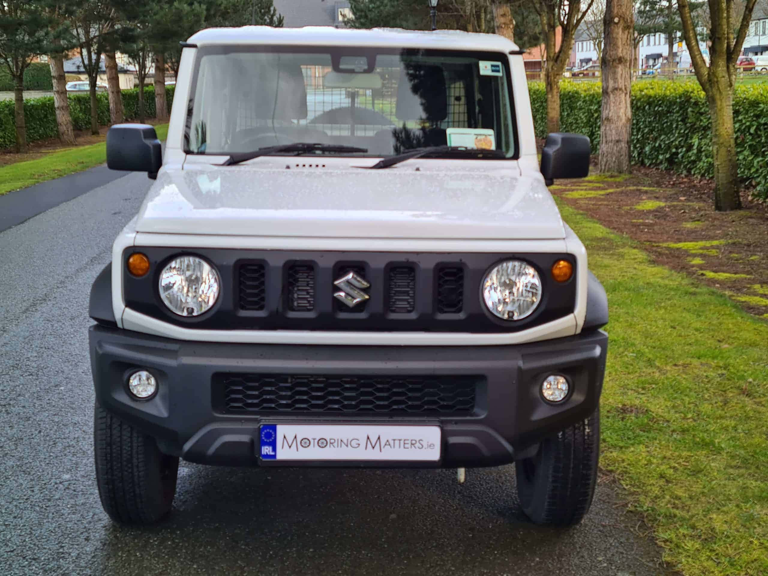 Suzuki Jimny 桌布 壁紙 — 一輛白色鈴木 Jimny SUV 停放在濕瀝青路上，兩旁有綠草和樹木。