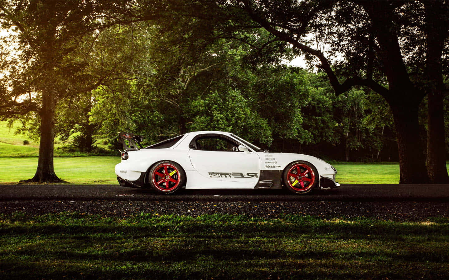  壁紙 — 一輛白色Mazda RX-7跑車，紅色輪圈，停泊在陽光普照的公園內，碎石路上，樹蔭之下。