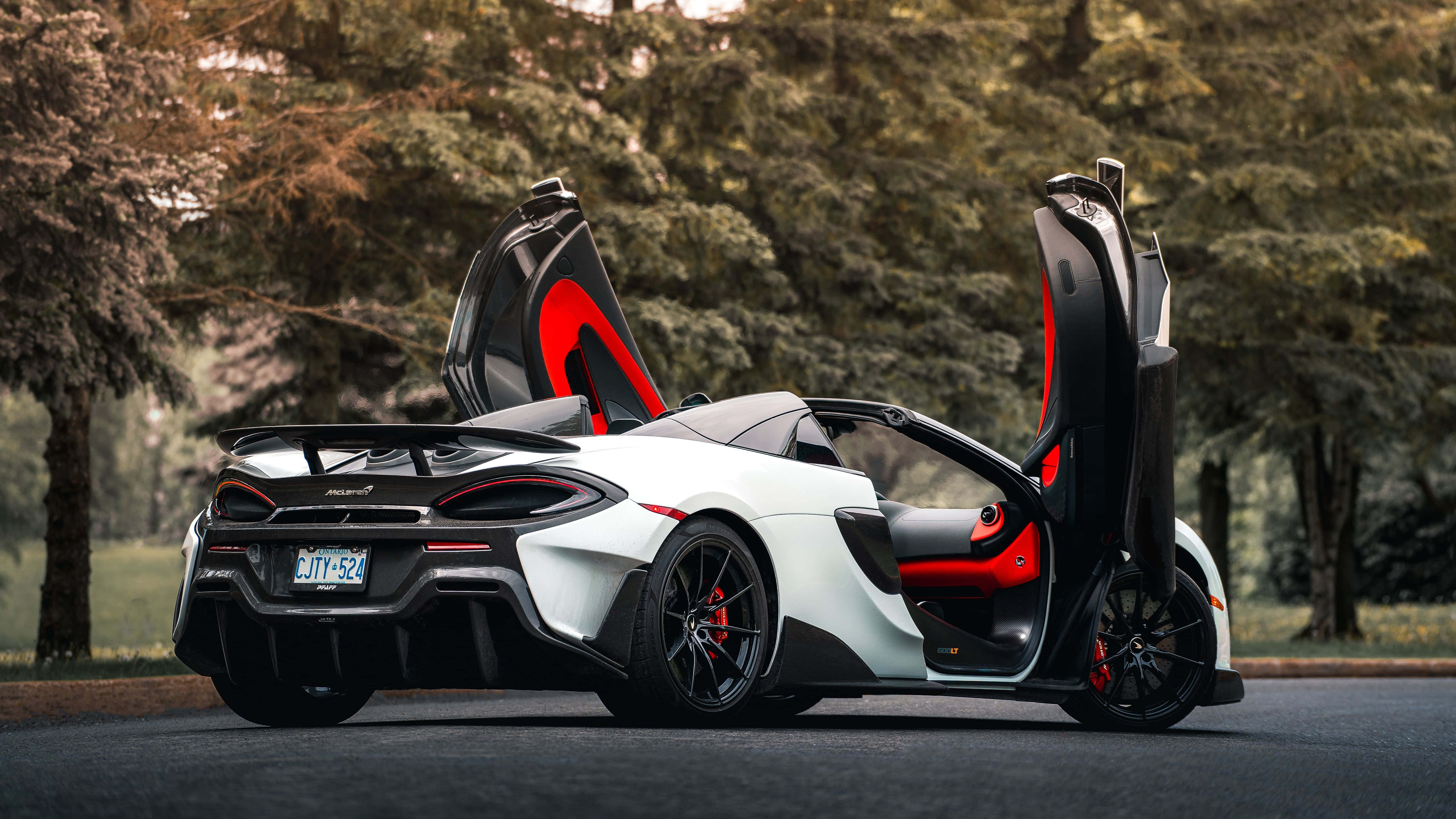 Mclaren 600lt 桌布 壁紙 — 一輛帶有紅色點綴和打開蝴蝶門的白色McLaren跑車停泊在林蔭大道上。