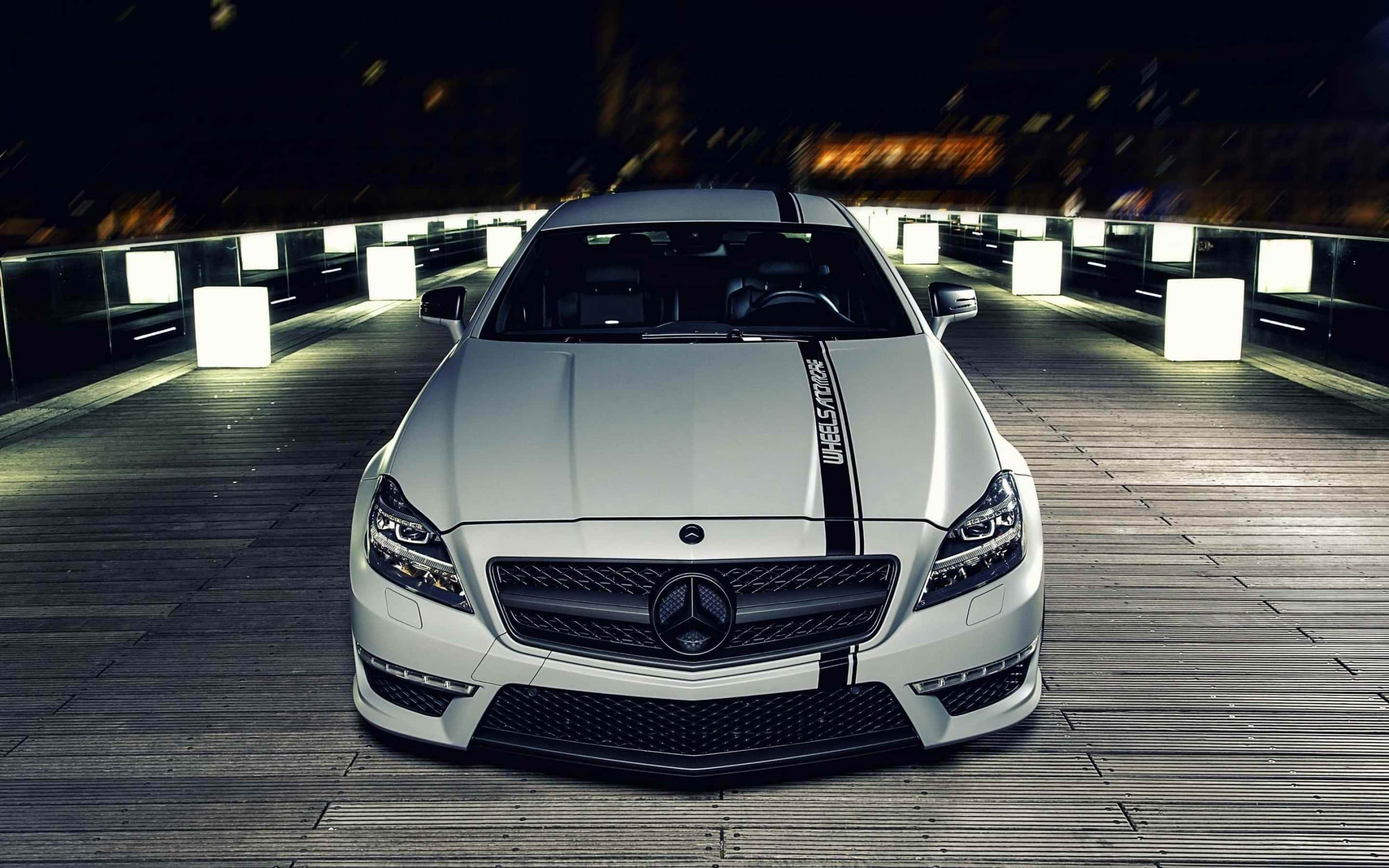 Mercedes Benz Cls-class 桌布 壁紙 — 一輛帶有黑色賽車條紋的時尚白色梅賽德斯-AMG跑車夜間停在木橋上，被發光的白色立方體和模糊的城市燈光照亮。