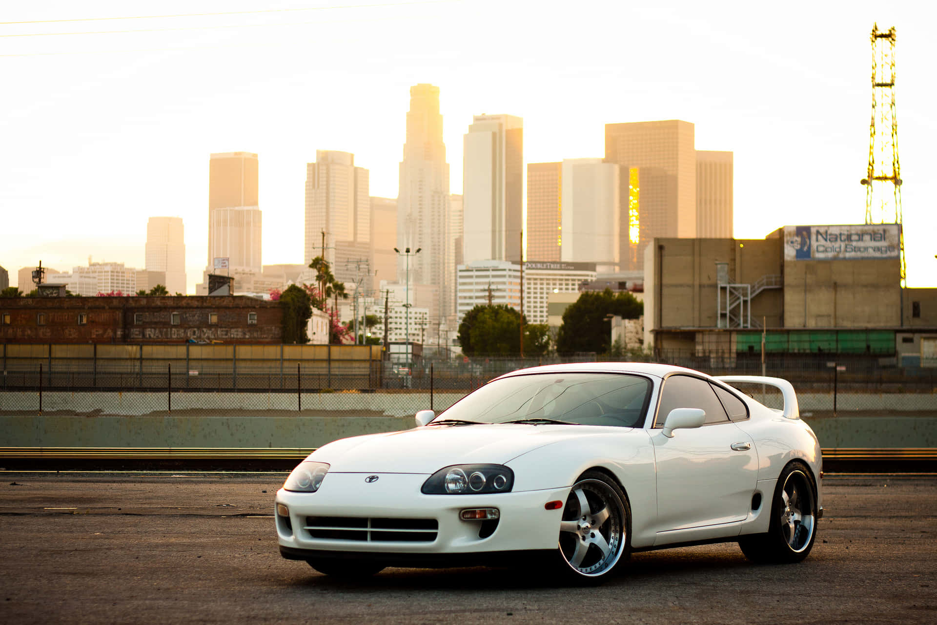 白色跑車停放在城市街道上，背景是夕陽下的城市天際線。 — Toyota Supra 壁紙