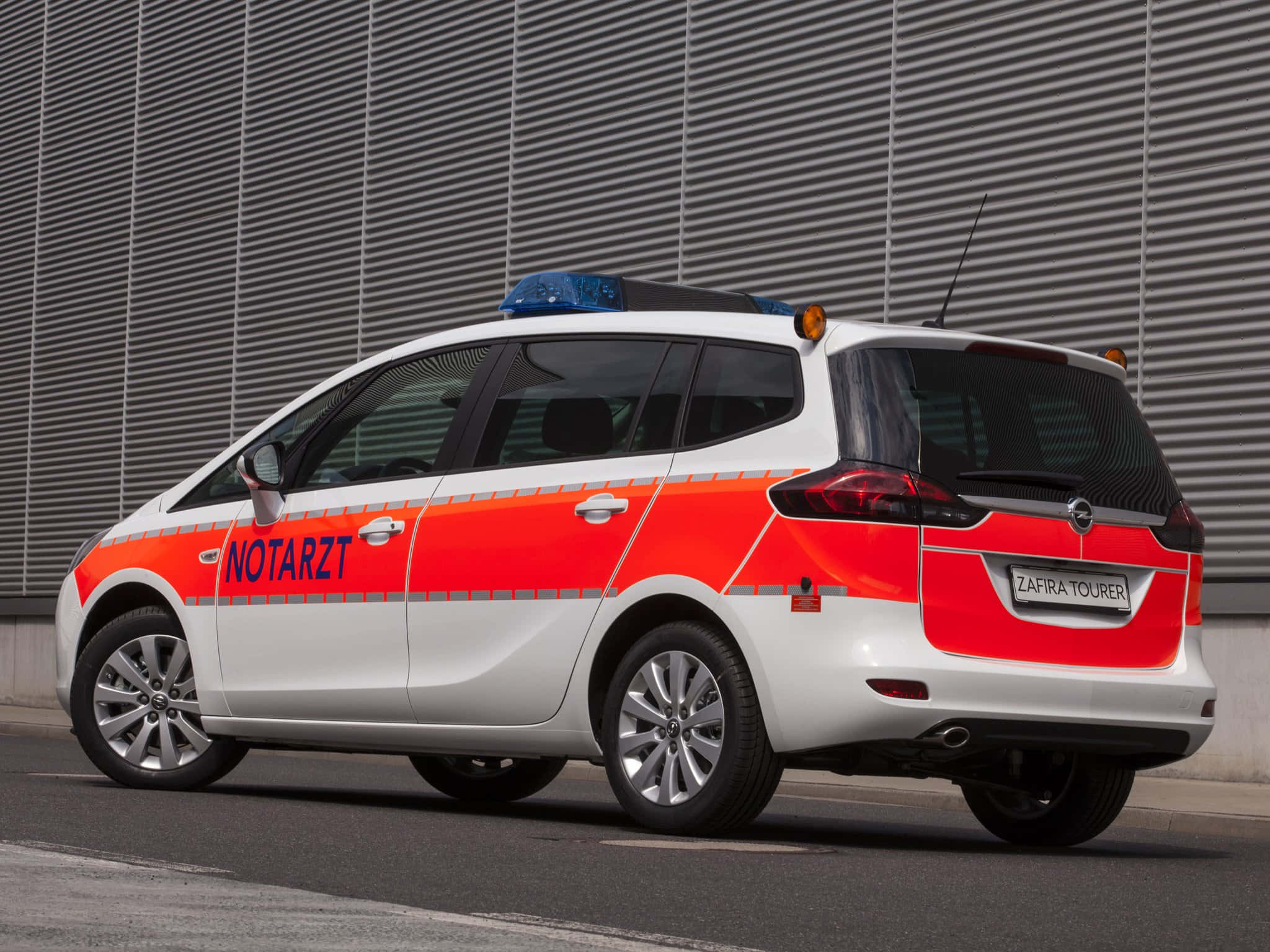 Opel Zafira 桌布 壁紙 — 一輛白色和橙色的德國緊急醫療服務車停靠在灰色波紋金屬牆旁。