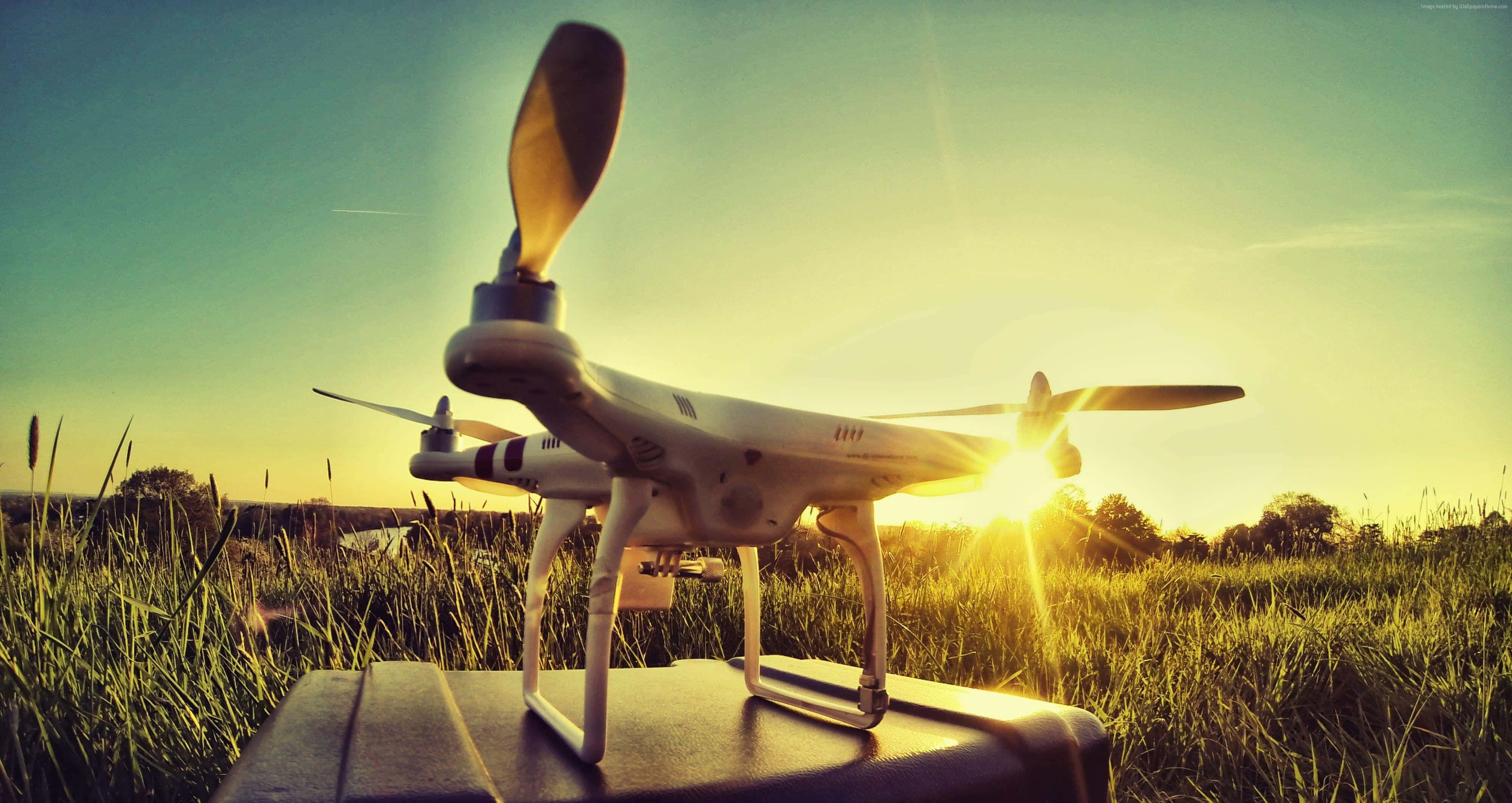 Drone Camera 桌布 壁紙 — 一架白色無人機放在草地上的一個箱子上，夕陽在它後面落下，散發出金色的光芒。