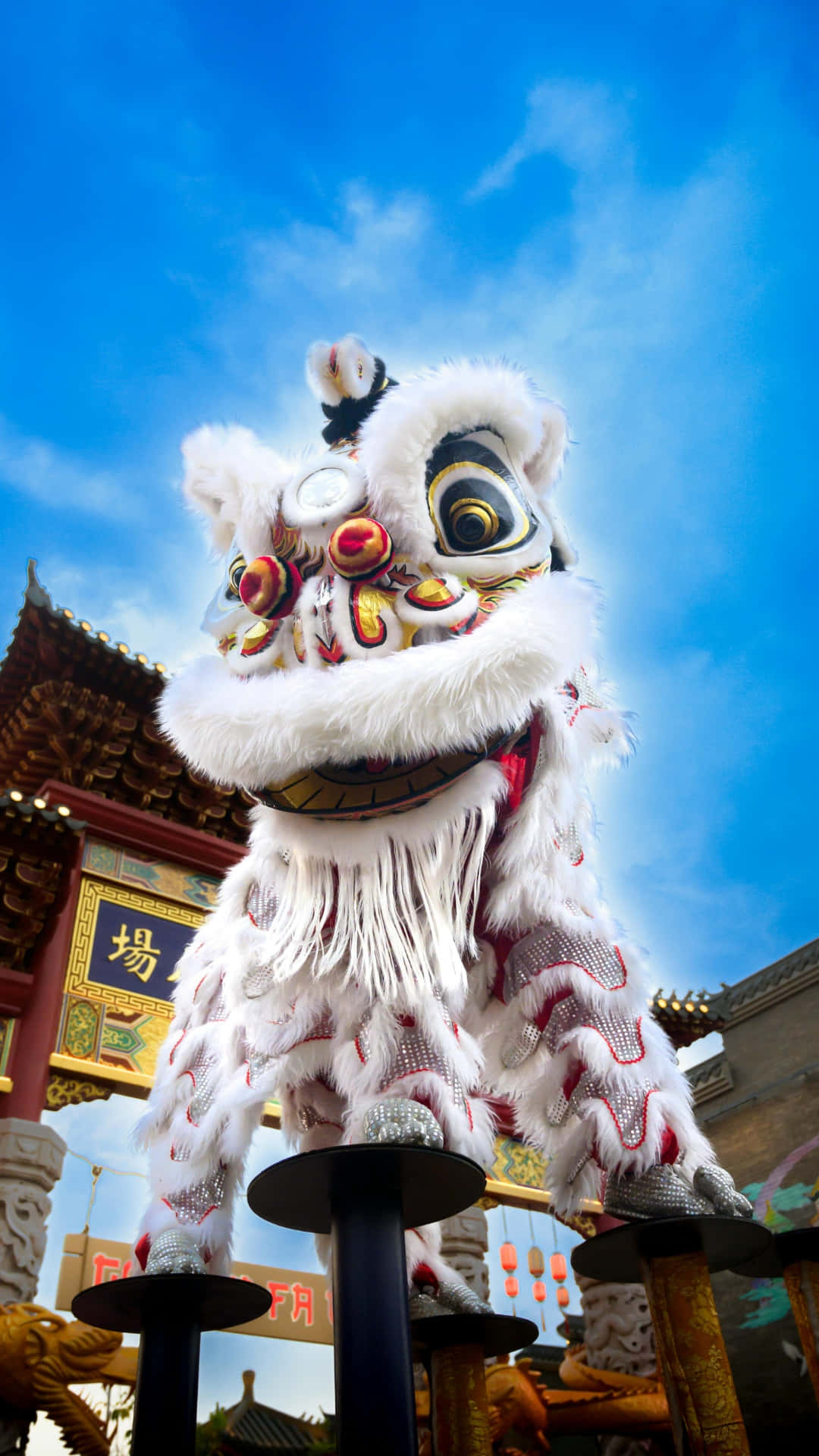  壁紙 — 一件充滿活力的白色舞獅服飾，帶有紅色點綴，在明亮的藍天下優雅地踩著高蹺，背景是傳統中式建築。