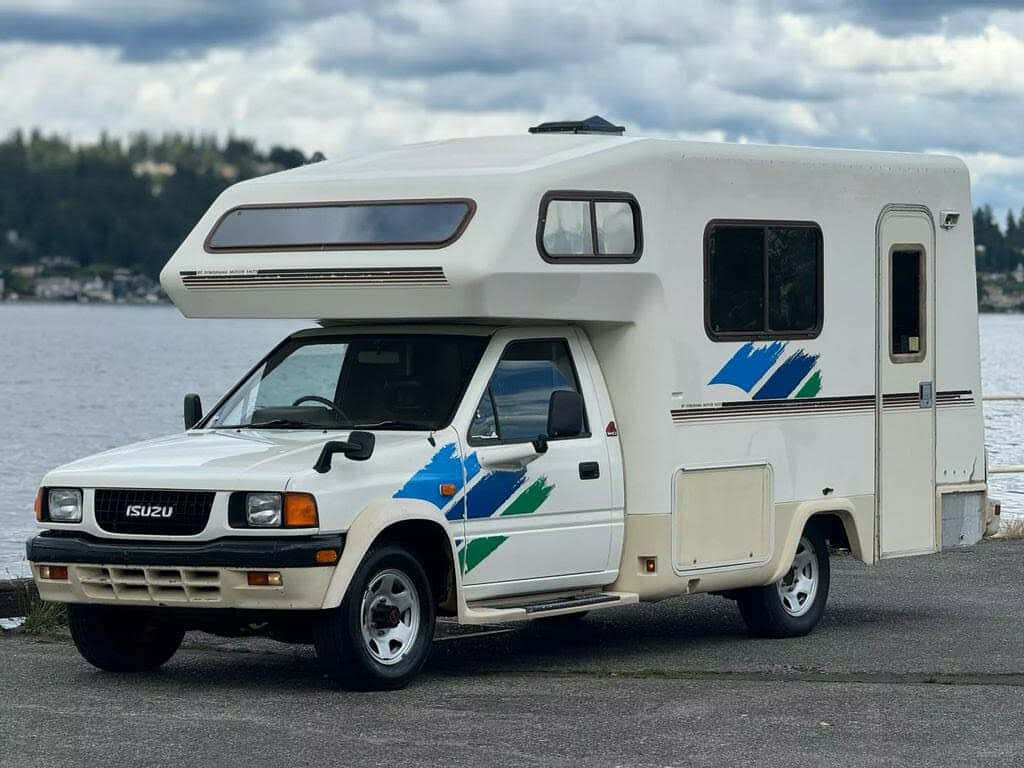  壁紙 — 一輛白色五十鈴露營車，帶有藍綠色貼花，停泊在平靜的湖邊，天空多雲。