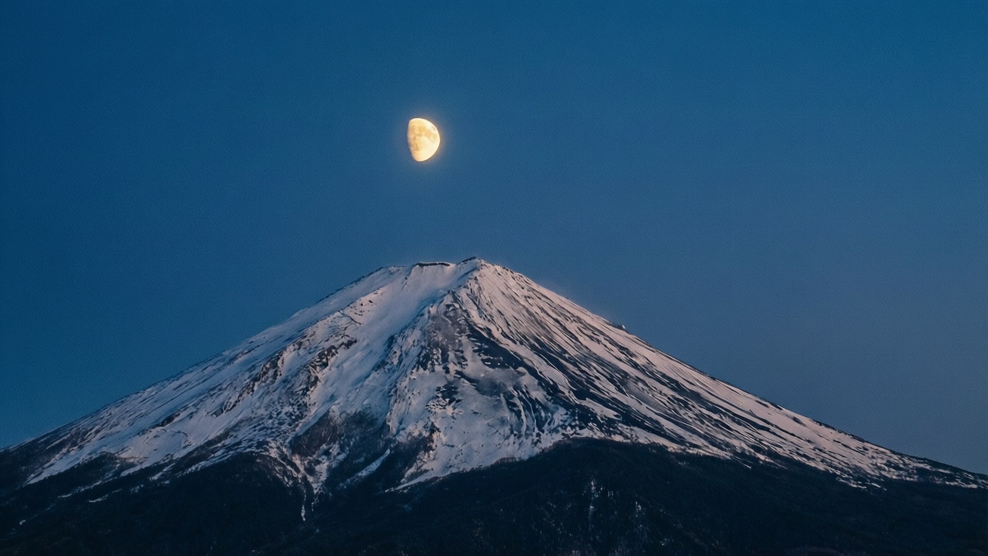 Moon over Peak 壁紙 — 白雪覆蓋的富士山在深藍色的天空下聳立，一輪發光的彎月直接位於其頂峰之上。