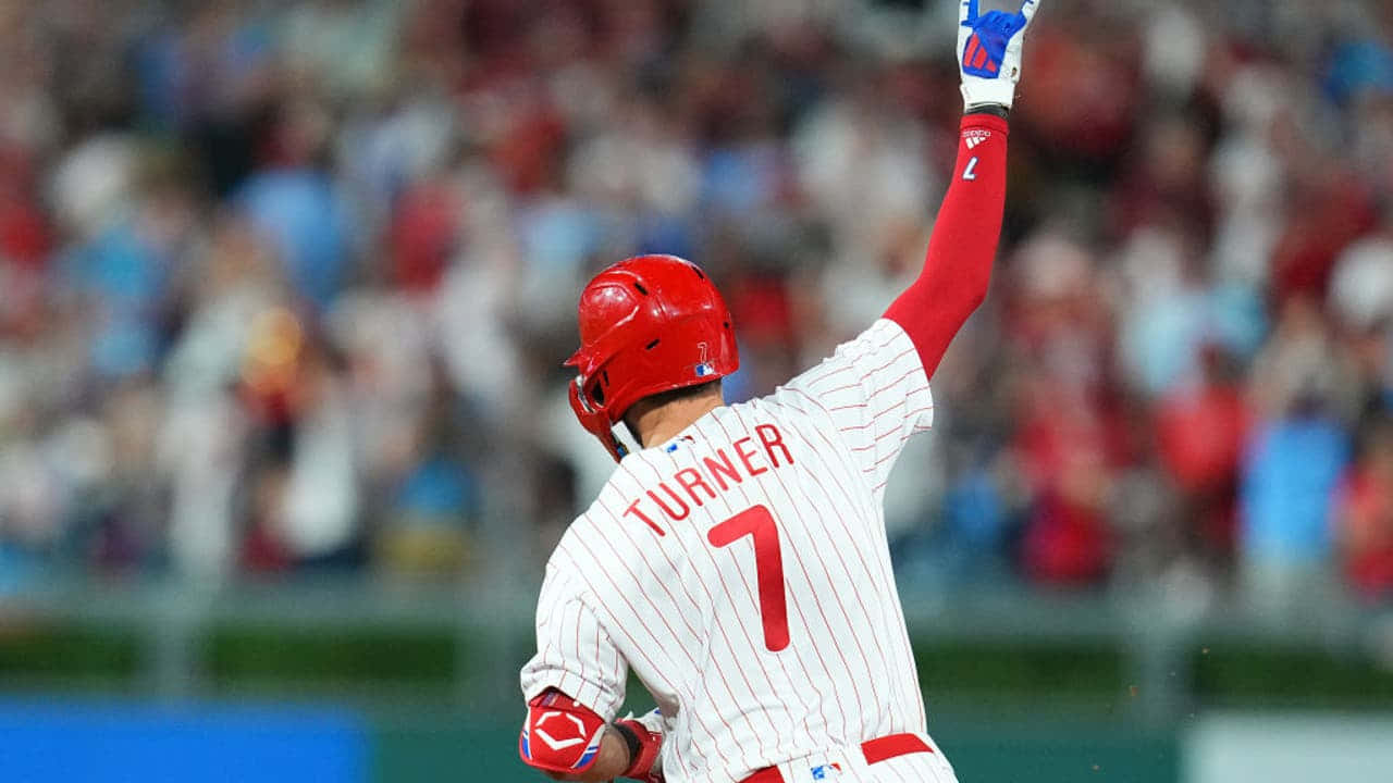 Trea Turner Phillies 桌布 壁紙 — 棒球運動員特納戴著紅色頭盔、身穿白色細條紋制服，在模糊的體育場人群中高舉戴手套的拳頭慶祝勝利。