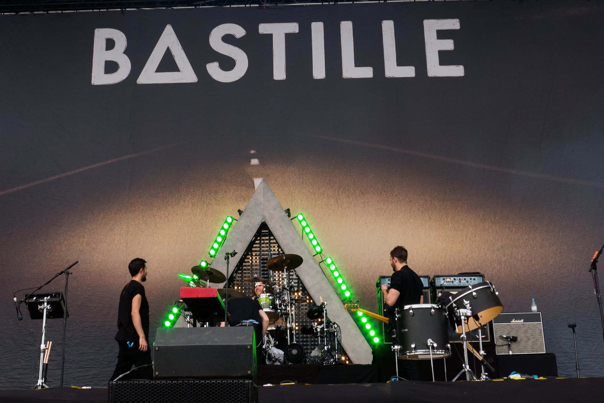 巴士底監獄 桌布 壁紙 — Bastille 樂團在舞台上現場表演，伴隨著發光的綠色燈光和一個大型三角形背景。