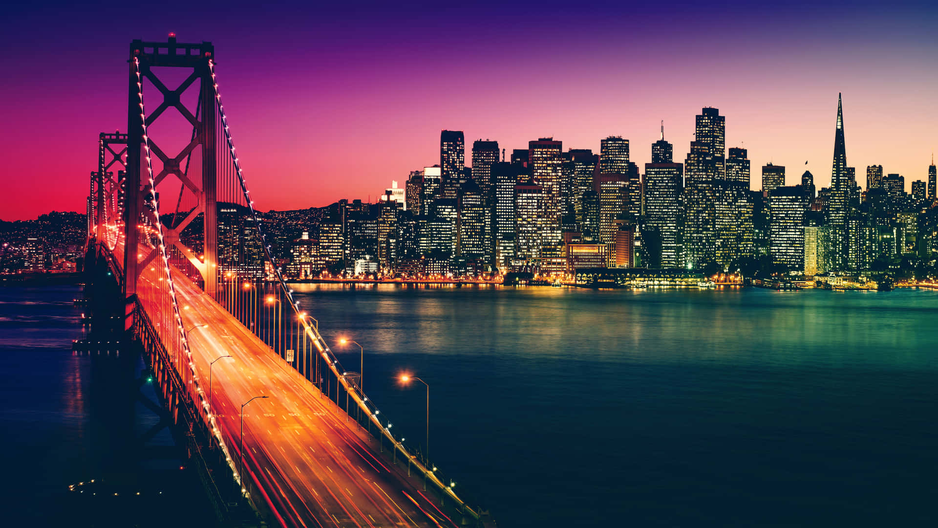  壁紙 — 海灣大橋在暮色中的 vibrant 景觀，車燈劃出光軌，燈火通明的舊金山天際線倒映在平靜的水面上。