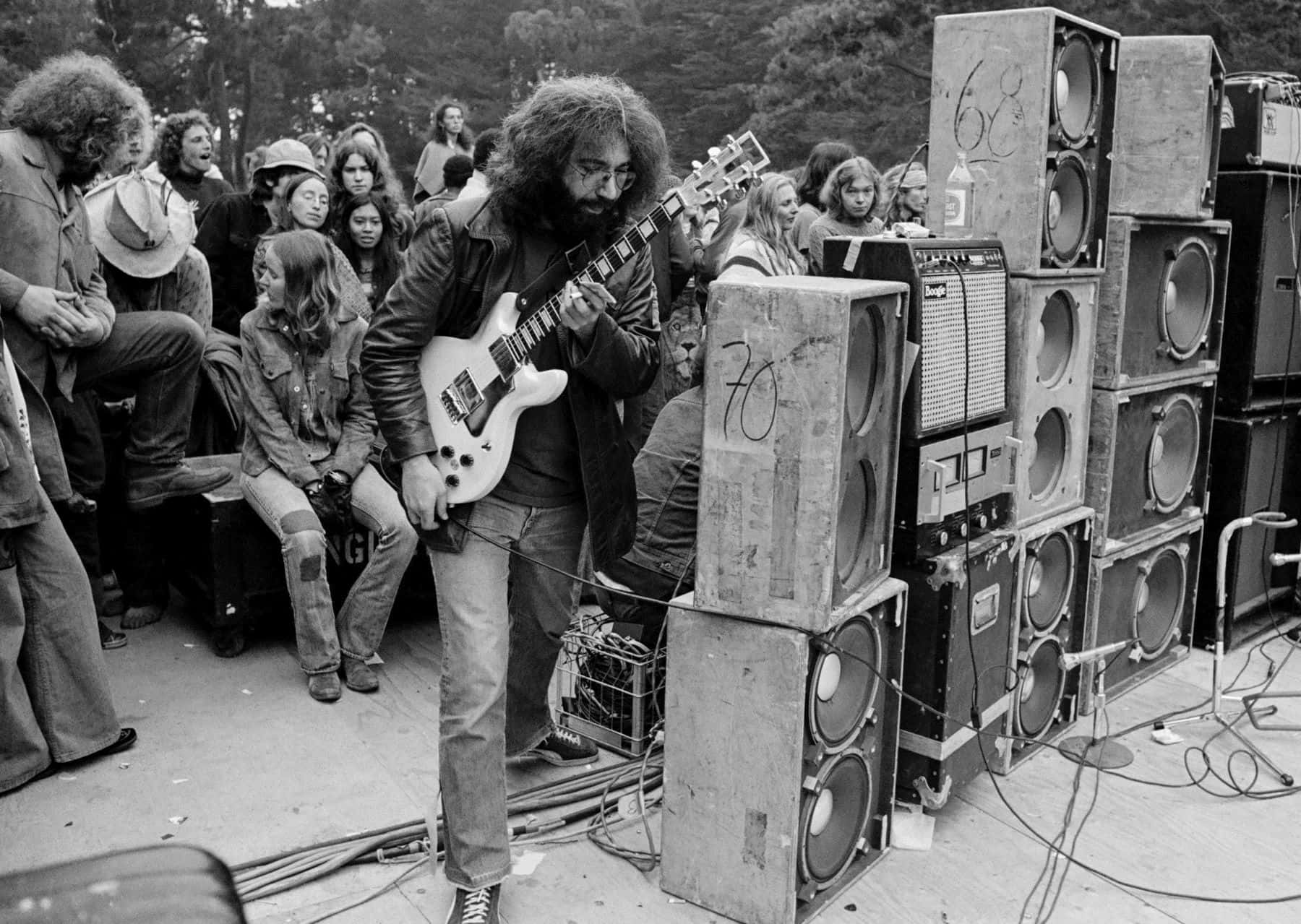 壁紙 — 一位留鬍鬚的吉他手在1970年代戶外音樂會的舞台上，被堆疊的擴音器和人群包圍，彈奏白色電吉他。