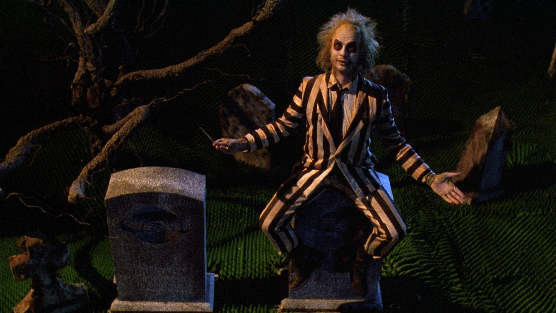 Beetlejuice 桌布 壁紙 — Beetlejuice 坐在墓碑上，在一個黑暗陰森的墓地裡，有扭曲的樹木和夜光下發光的綠色草地。