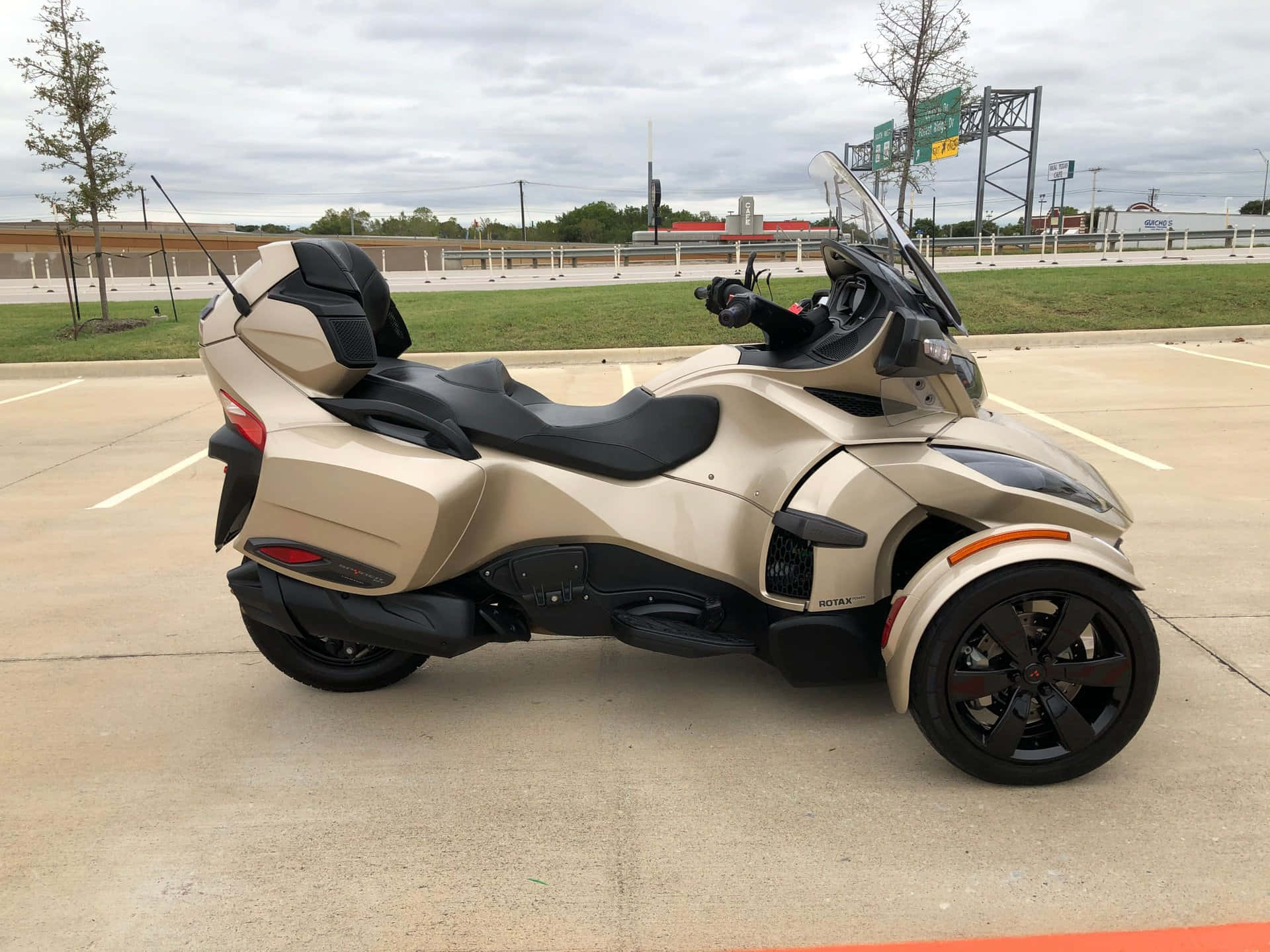  壁紙 — 一輛米色和黑色的三輪Can-Am Spyder RT停放在空曠的混凝土場地上，天空多雲。