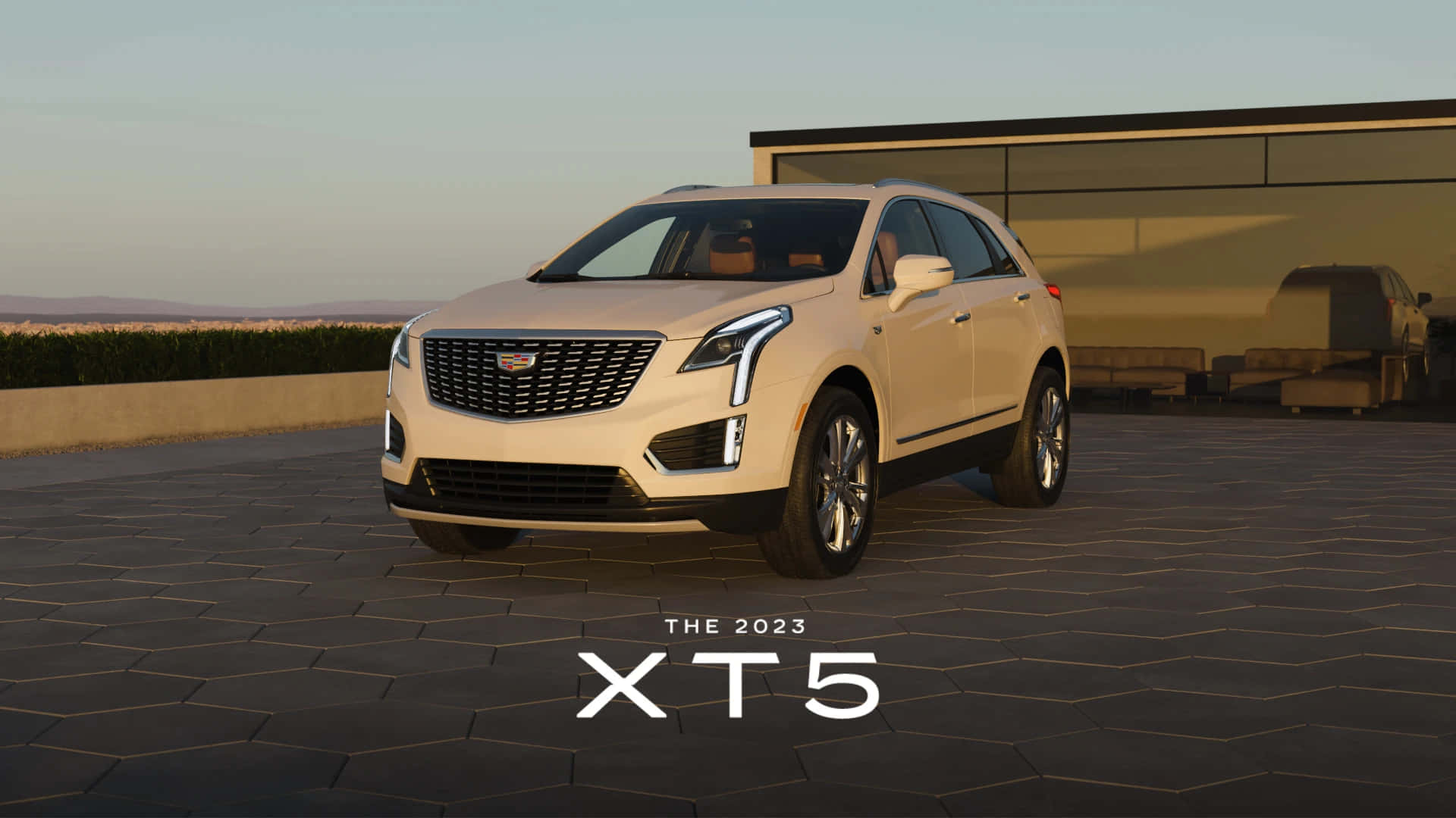 Cadillac Xt5 桌布 壁紙 — 一輛流線型的米色2023年凱迪拉克XT5 SUV停放在六角形石頭庭院上，旁邊有現代房屋和遠處的山丘，背景是柔和的金色日落天空。