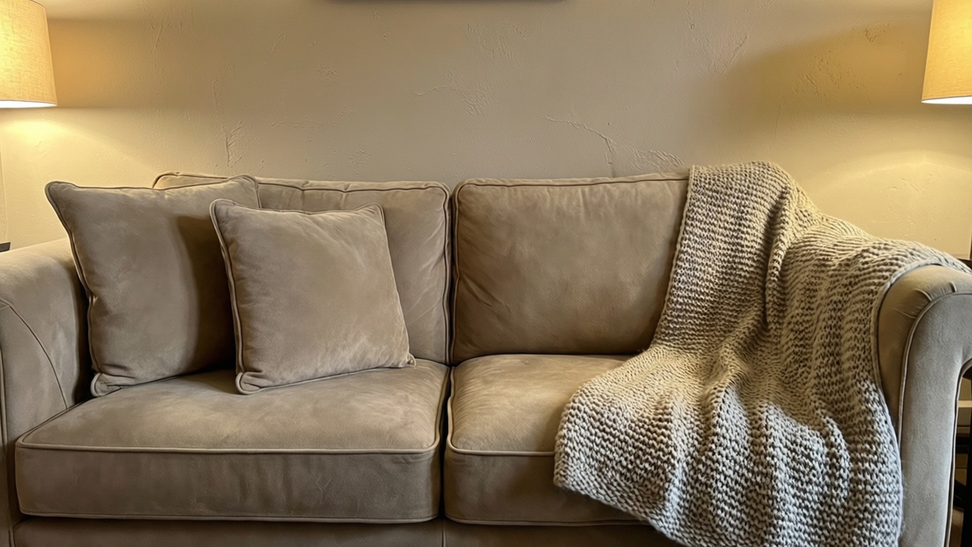 Beige Suede Sofa 壁紙 — 一張米色天鵝絨沙發，搭配相襯的抱枕，以及一條厚針織毛毯搭在其中一邊扶手上，在溫暖的燈光下。