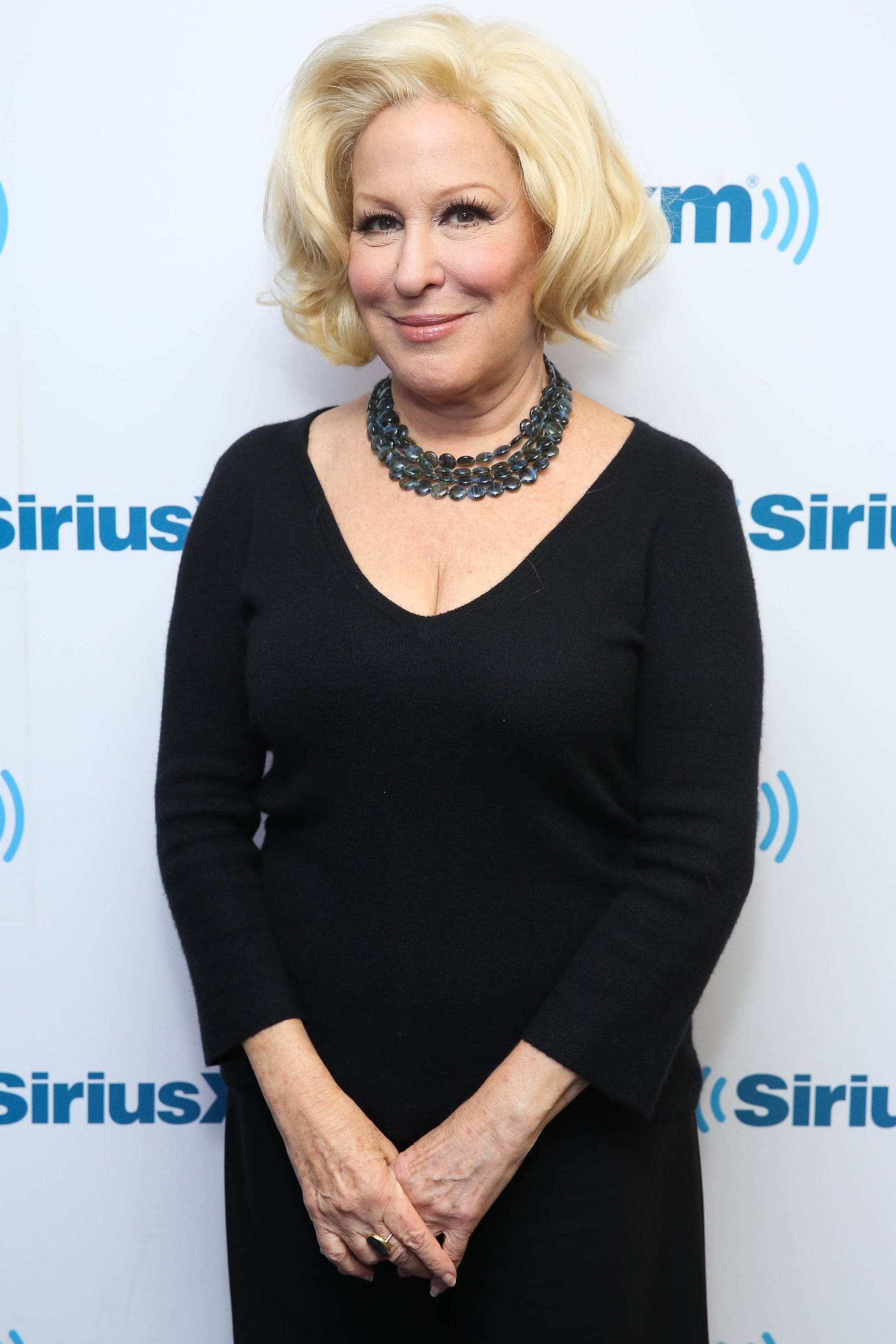 Singer 桌布 壁紙 — Bette Midler 身穿黑色連衣裙和層疊項鍊，在SiriusXM品牌背景前溫暖地微笑。
