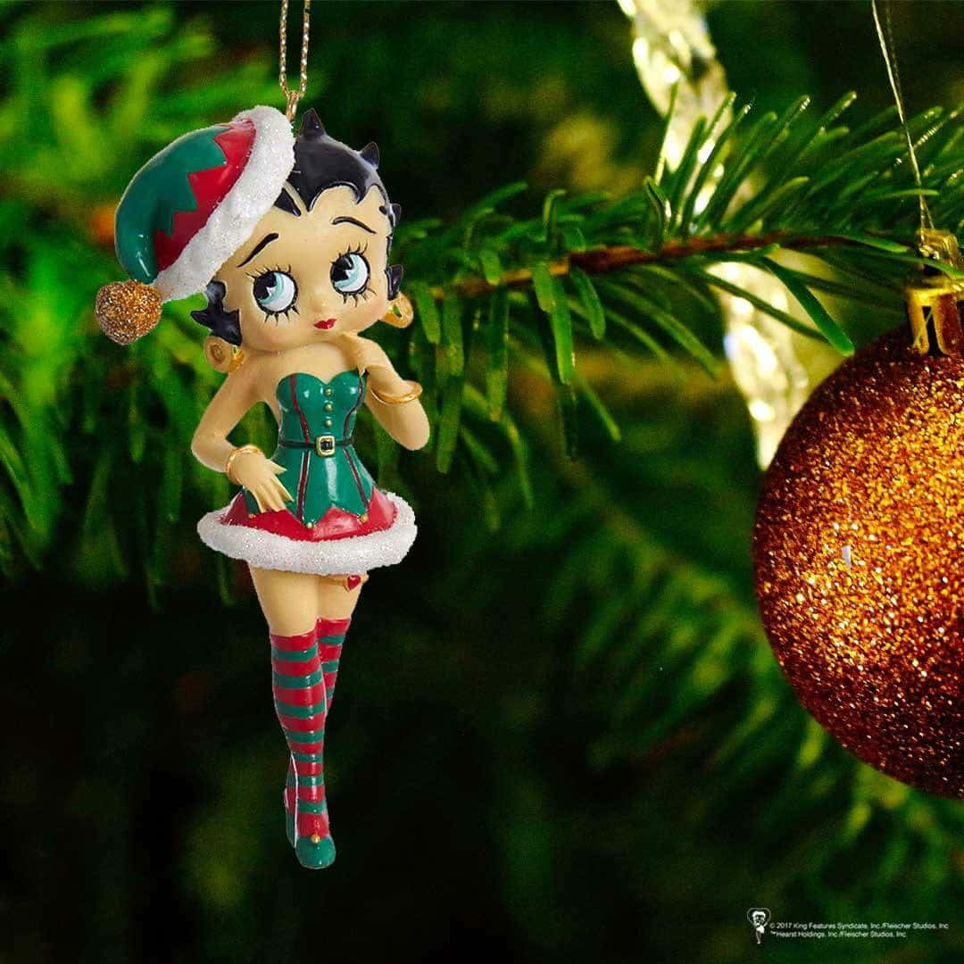 Betty Boop Christmas 桌布 壁紙 — Betty Boop 聖誕裝飾，身穿綠色和紅色精靈服，掛在松樹上，旁邊有一個閃閃發光的金色聖誕球。