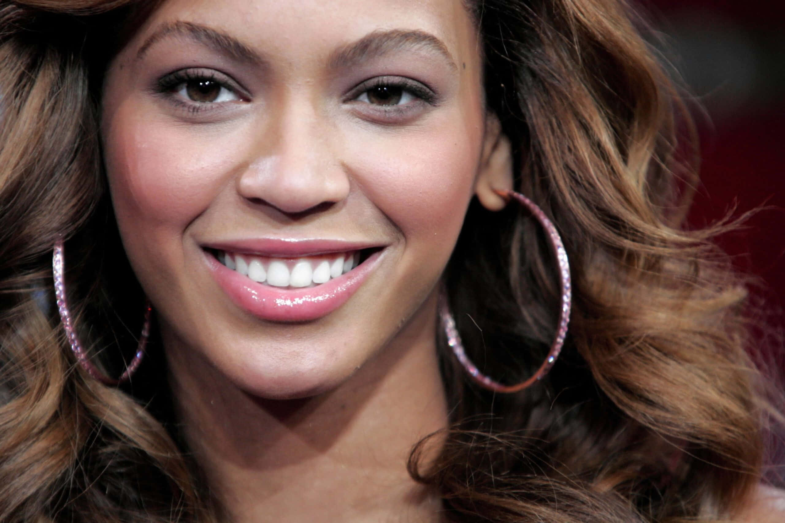 Smile 桌布 壁紙 — Beyoncé 在模糊的紅色背景前，戴著閃閃發光的圈形耳環和一頭濃密的棕色捲髮，燦爛地微笑。