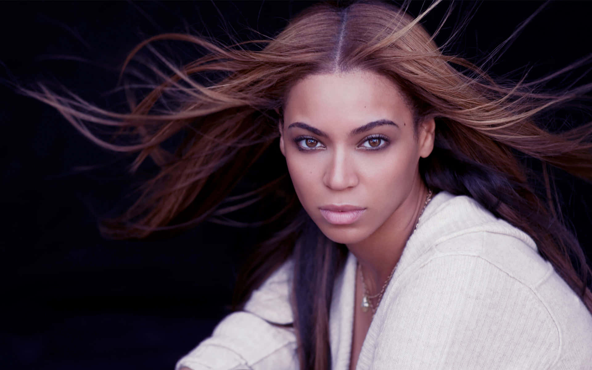 碧昂絲 桌布 壁紙 — Beyoncé 在深色背景下，棕色頭髮隨風飄揚，穿著白色連帽衫，凝視著。