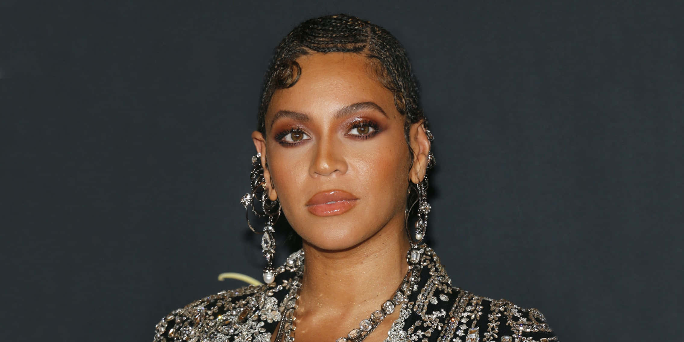 碧昂絲 桌布 壁紙 — Beyoncé 在深色背景下，身穿閃亮黑色服裝，搭配精緻辮子髮型和垂墜耳環擺姿勢。