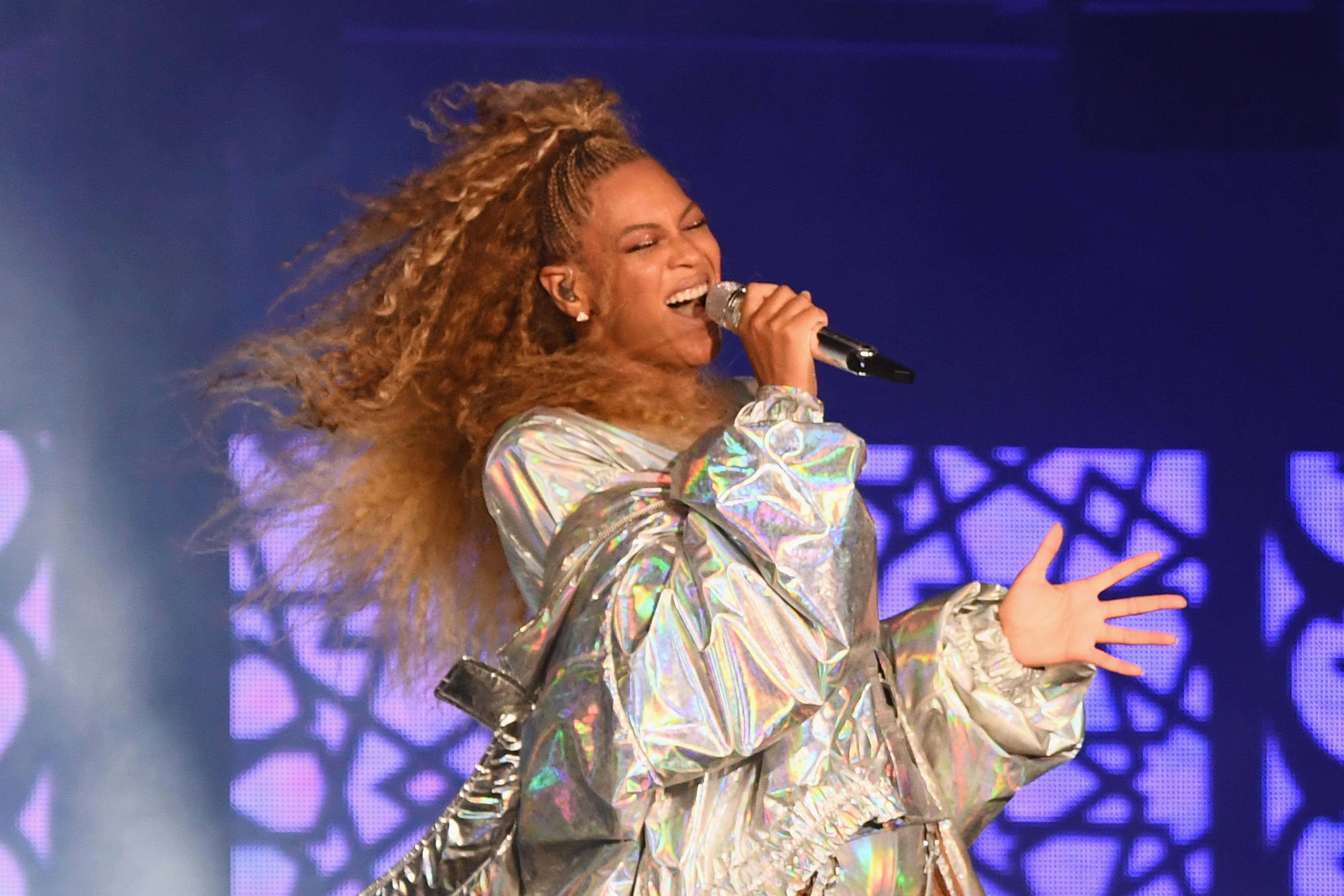 碧昂絲 桌布 壁紙 — Beyoncé 在紫色圖案背景前，身穿閃爍銀色服裝，長髮飄逸，在舞台上熱情表演。