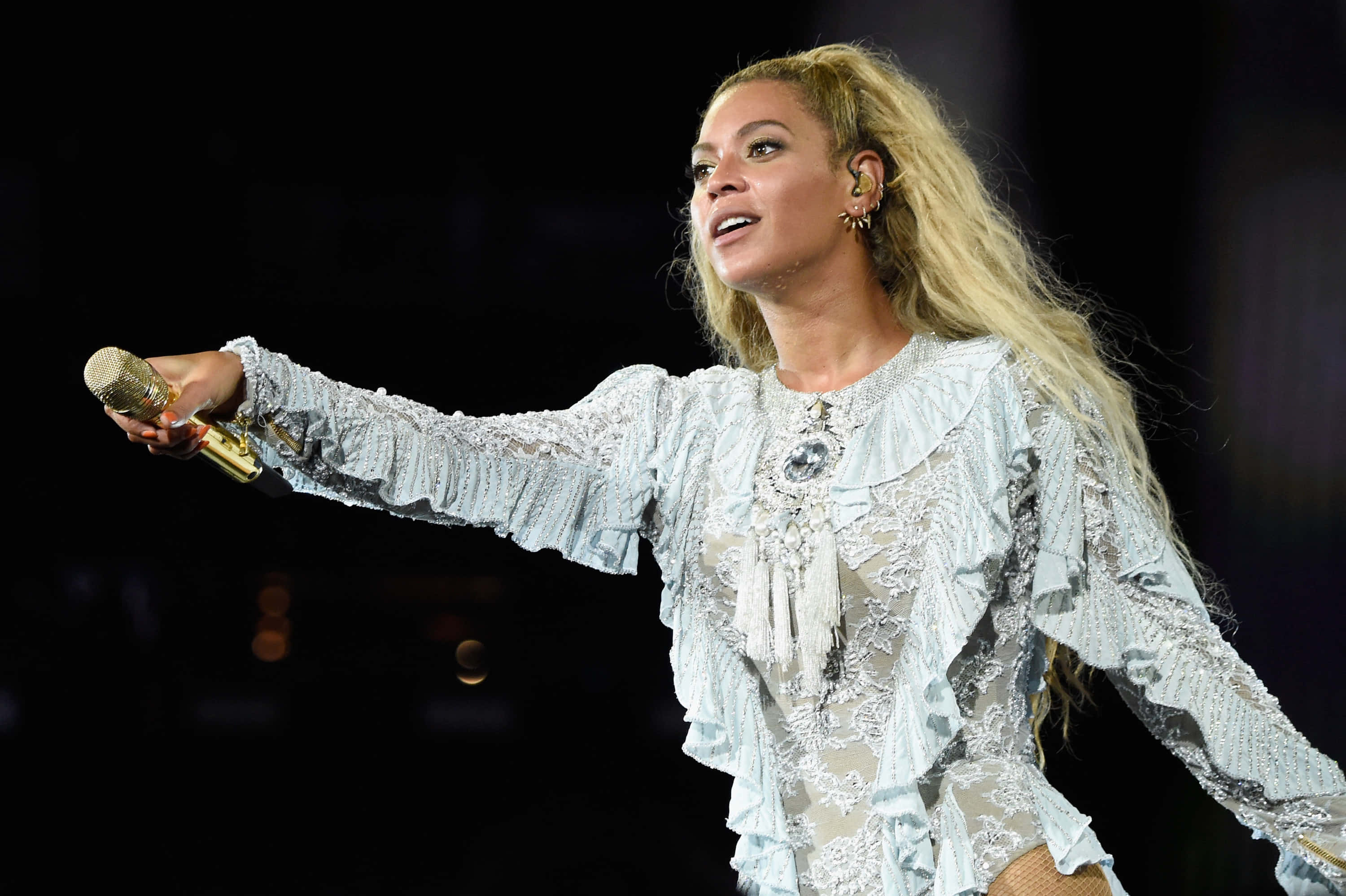 碧昂絲 桌布 壁紙 — Beyoncé 在舞台上穿著精緻的淺藍色蕾絲服裝，手持金色麥克風，在舞台燈光下表演。