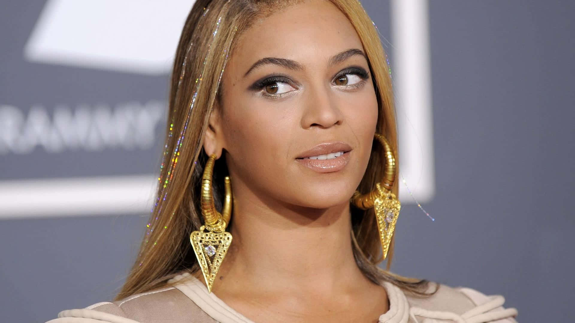 碧昂絲 桌布 壁紙 — Beyoncé 在葛萊美獎上佩戴金色耳環和米色服裝，帶有閃亮的頭髮裝飾。