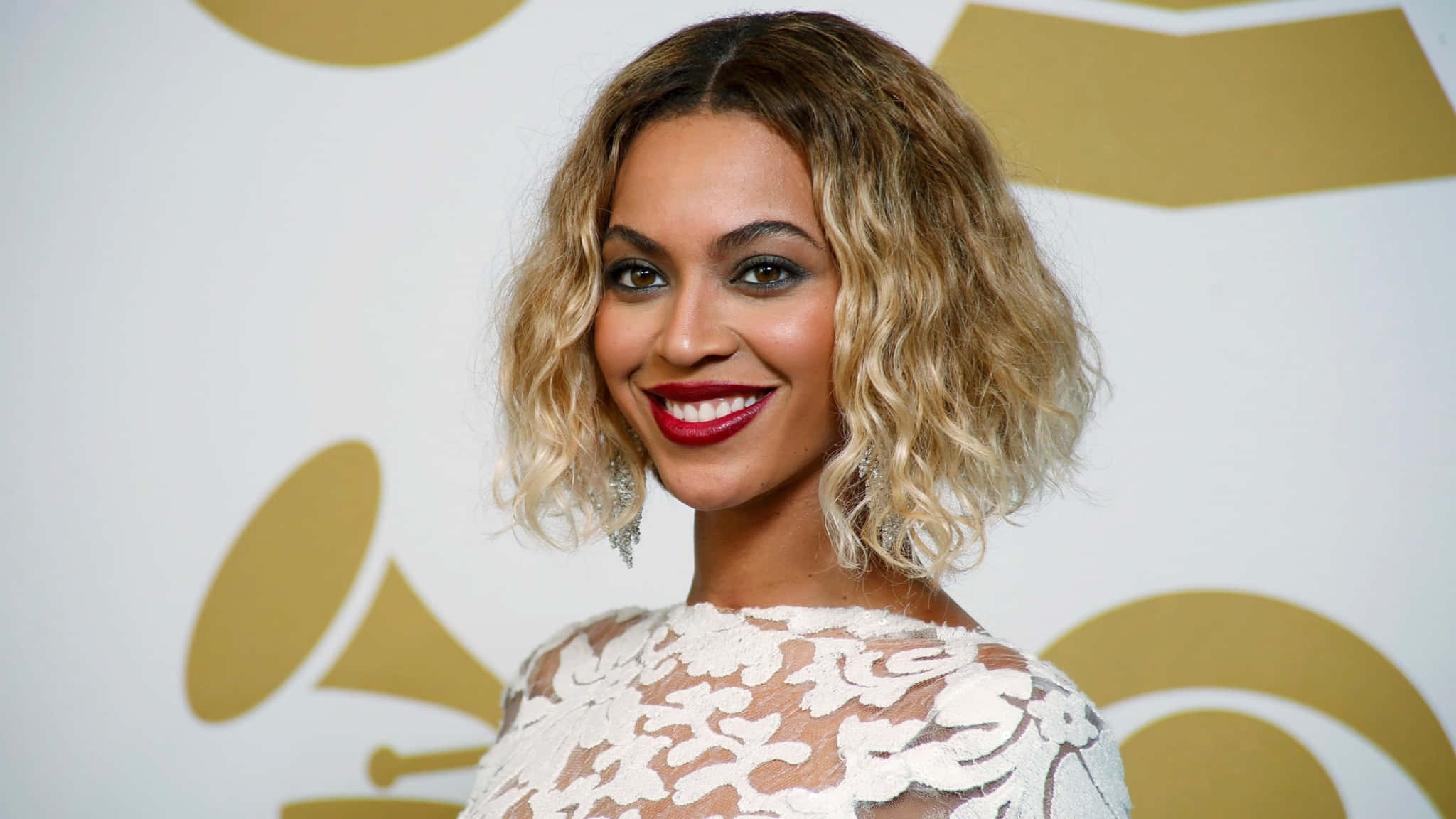 碧昂絲 桌布 壁紙 — Beyoncé 在金色葛萊美標誌背景下穿著白色蕾絲連衣裙燦爛微笑。
