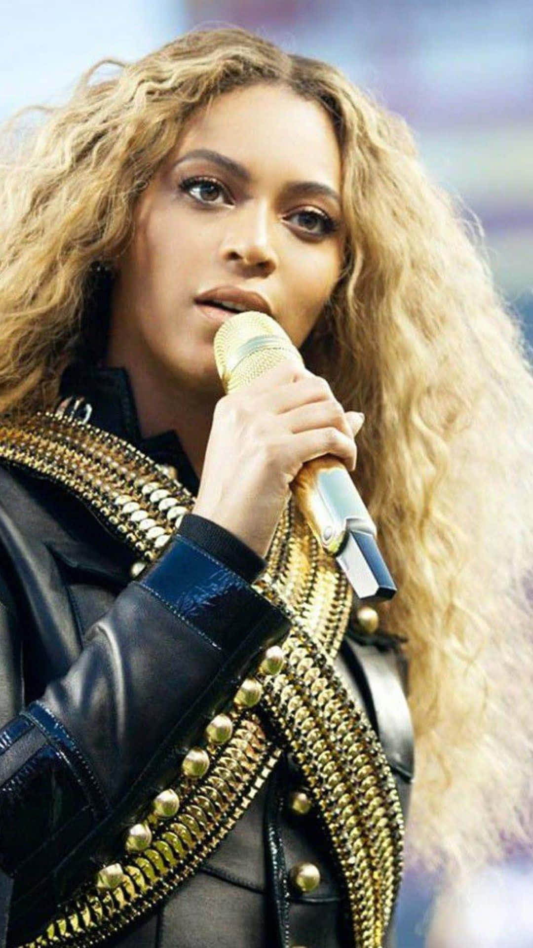 碧昂絲 桌布 壁紙 — Beyoncé 專注地表演，手持金色麥克風，身穿飾有金色鏈條的黑色皮夾克。