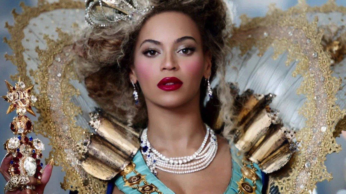 碧昂絲 桌布 壁紙 — Beyoncé 戴著華麗的金色王冠和精緻珠寶，手持鑲有寶石的權杖，背景是柔和的藍色。