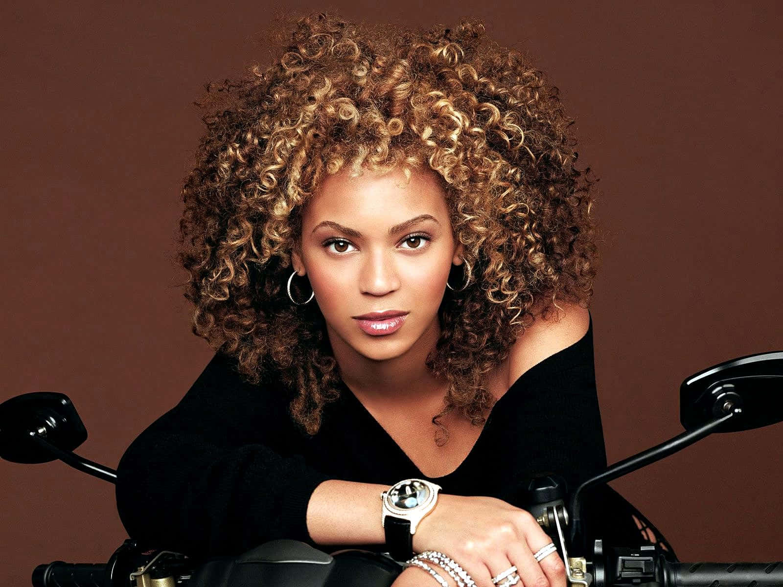 碧昂絲 桌布 壁紙 — Beyoncé 擁有一頭濃密捲髮，自信地靠在一輛摩托車上，背景是豐富的棕色。