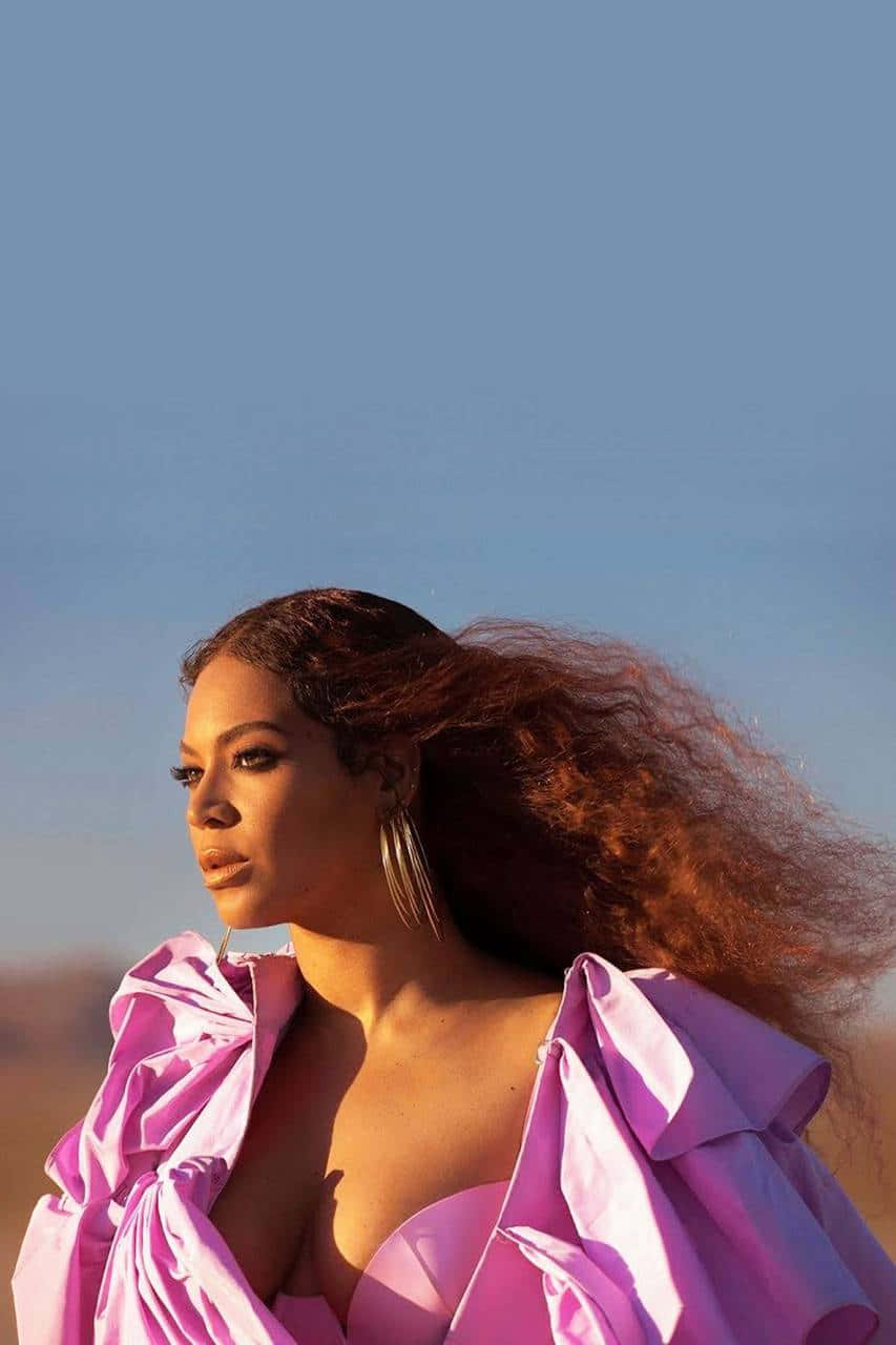 碧昂絲 桌布 壁紙 — Beyoncé 站在沙漠景觀中，穿著充滿活力的粉紅色荷葉邊連衣裙，風吹過她長長的捲髮，在清澈的藍天下。