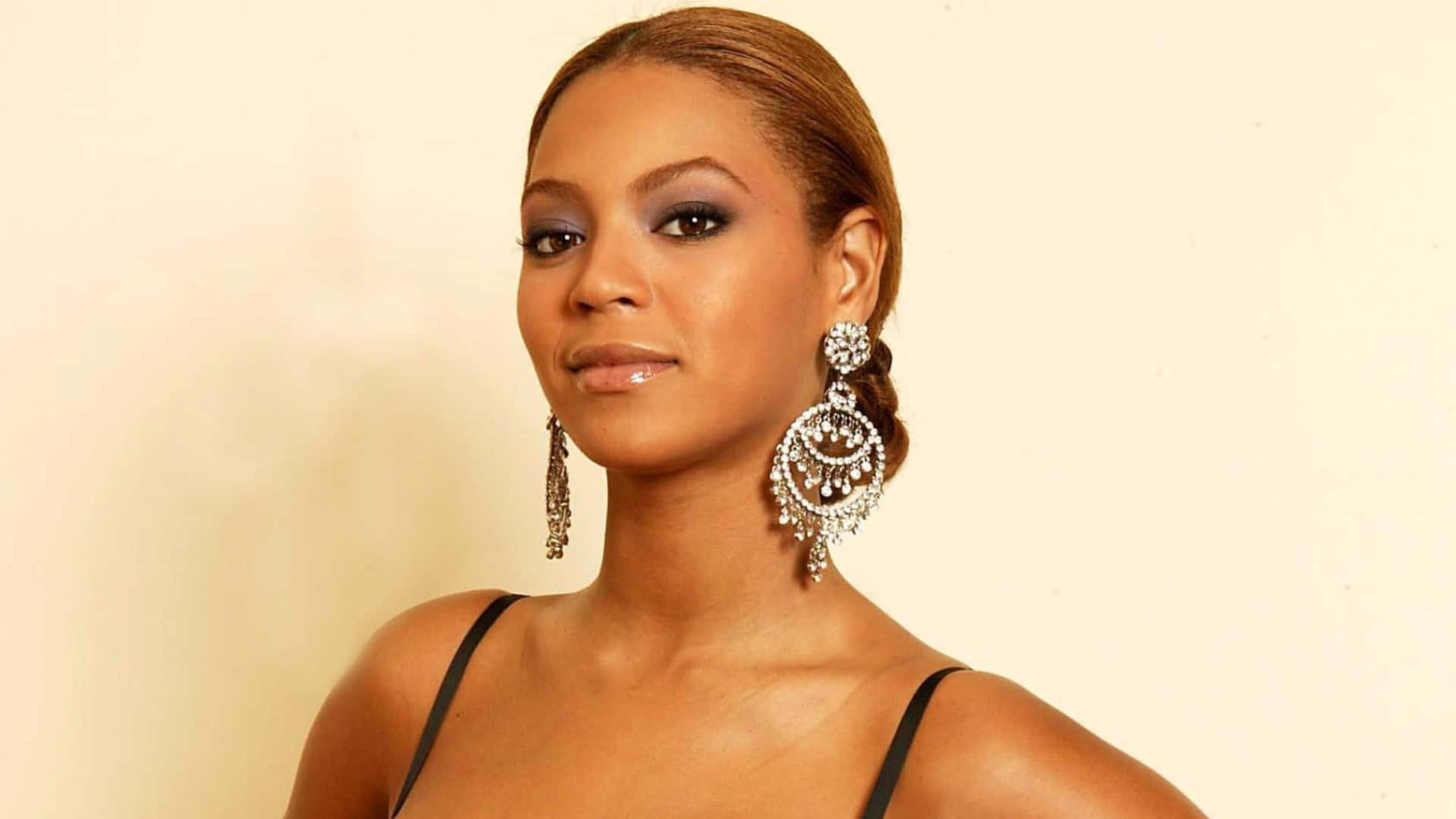 碧昂絲 桌布 壁紙 — Beyoncé 自信地擺姿勢，身穿黑色上衣，佩戴精緻的鑽石耳環，背景是柔和的米色。