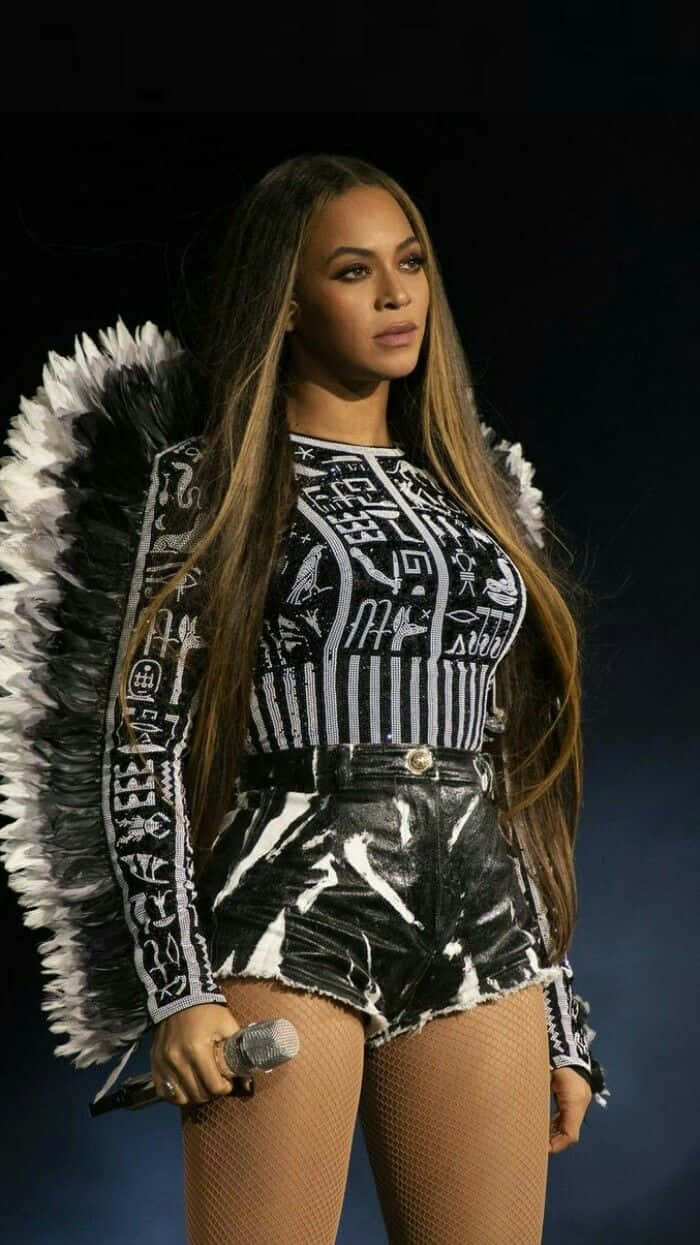 碧昂絲 桌布 壁紙 — Beyoncé 自信地站在舞台上，身穿帶有象形文字圖案的黑白羽毛天使服裝，手持麥克風。