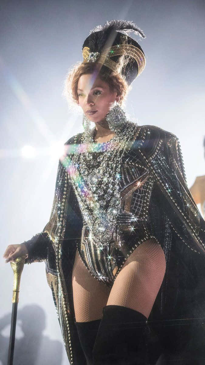 碧昂絲 桌布 壁紙 — Beyoncé 身穿閃耀的黑金珠寶緊身連衣褲，頭戴羽毛頭飾，手持手杖，被明亮的舞台燈光照亮。