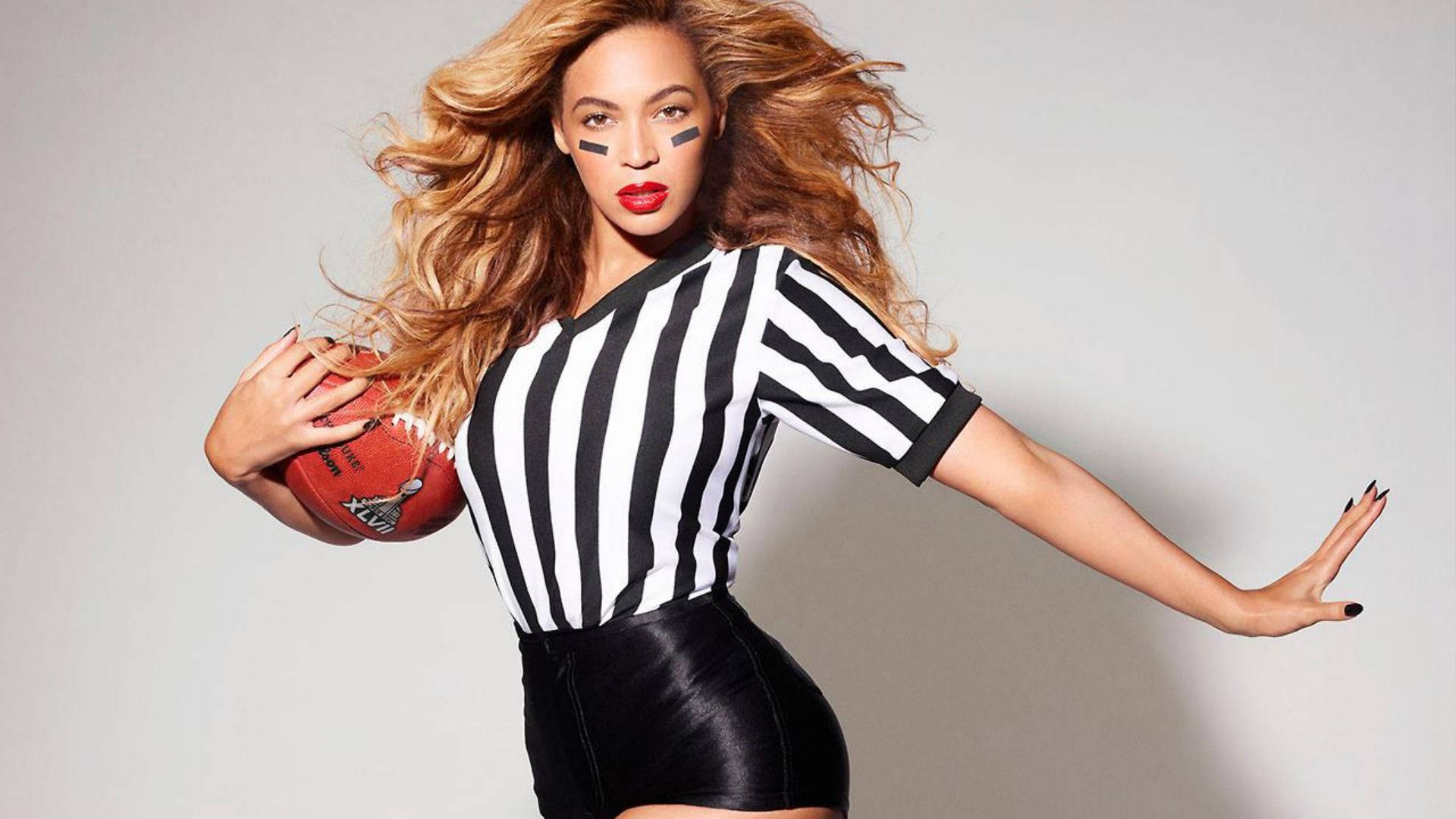 碧昂絲 桌布 壁紙 — Beyoncé 身穿黑白條紋裁判衫，手持美式足球，在純灰色背景前動感擺姿勢。