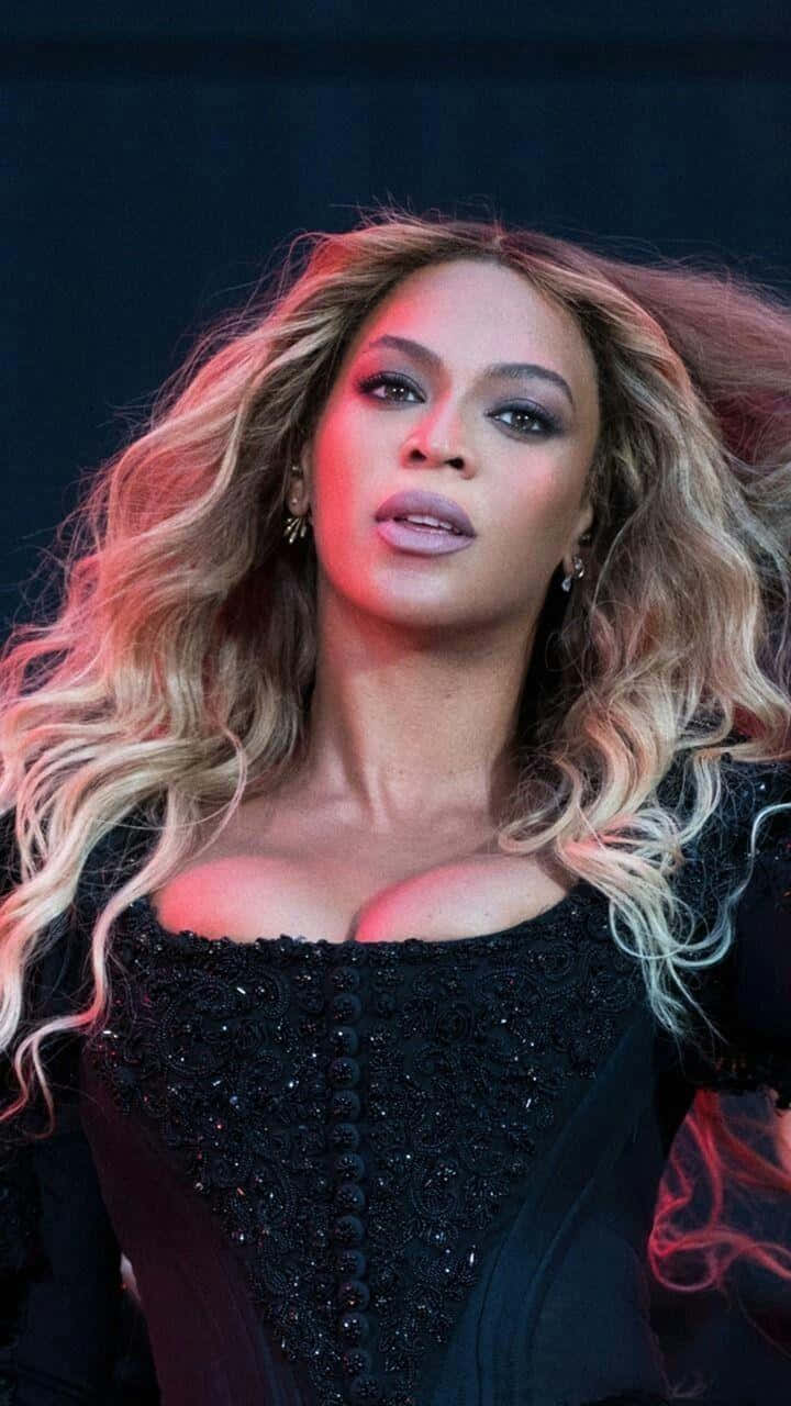 碧昂絲 桌布 壁紙 — Beyonce 一頭飄逸金髮，在紅色燈光下，身穿黑色鑲飾上衣，在深色背景中凝視。