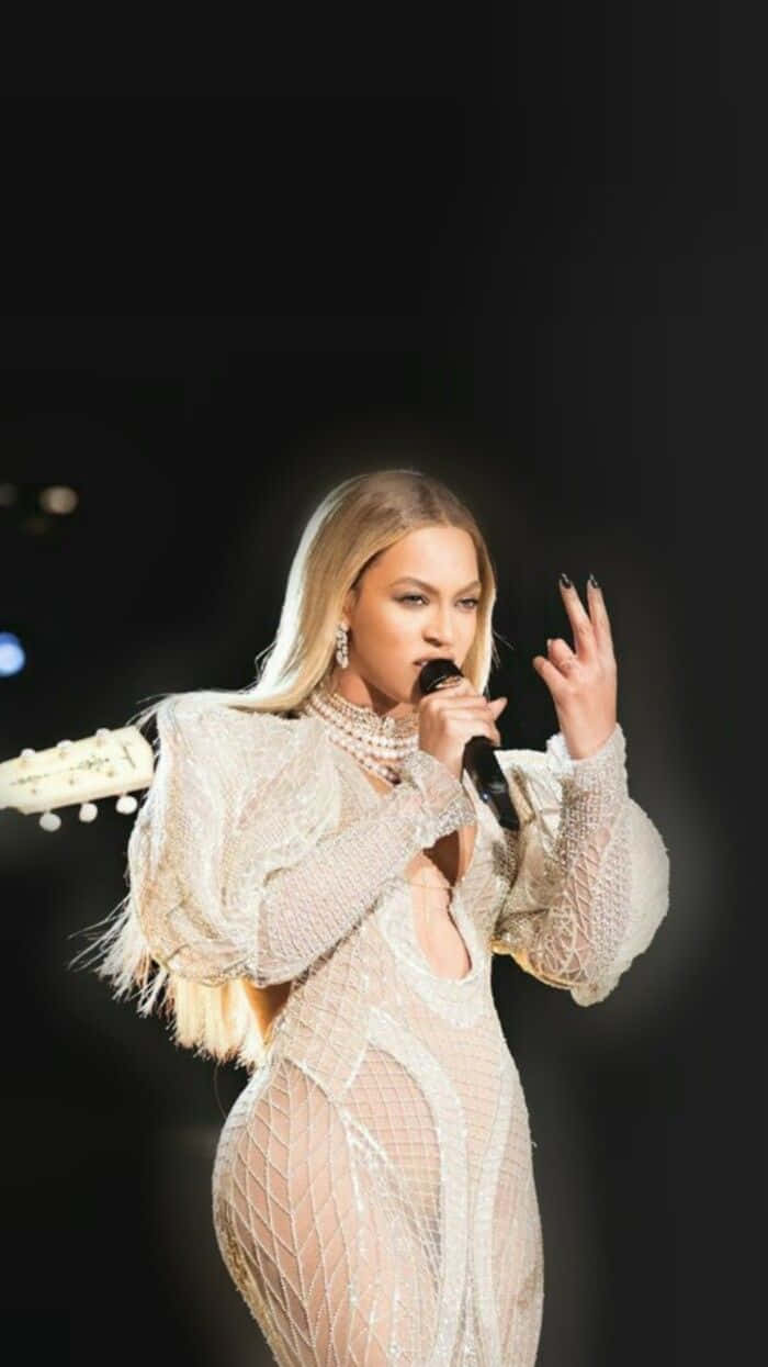 碧昂絲 桌布 壁紙 — Beyonce 在舞台上穿著閃閃發光的白色網狀長裙表演，手持麥克風，背後可見吉他帶。