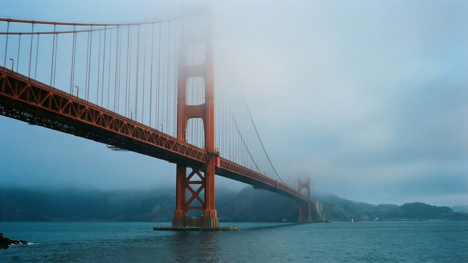 Golden Gate in Fog 壁紙 — 標誌性的橙色金門大橋從濃霧中浮現，下方是平靜的藍色海水，天空多雲。