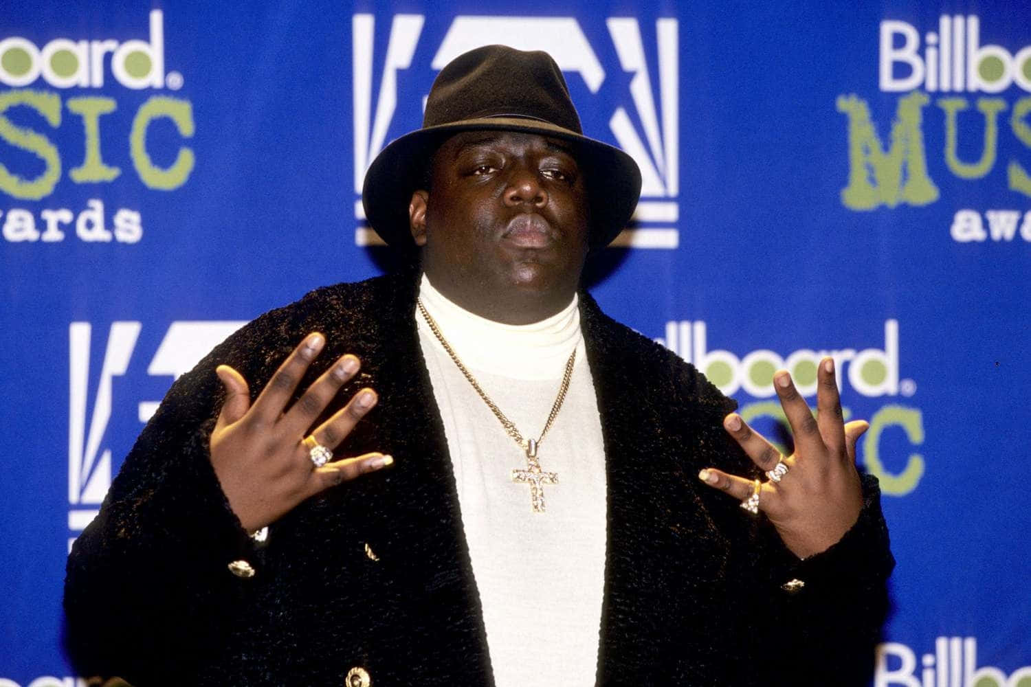  壁紙 — Biggie Smalls 在Billboard音樂獎上自信地擺姿勢，戴著黑色軟呢帽和金色珠寶，背景為藍色品牌背景。